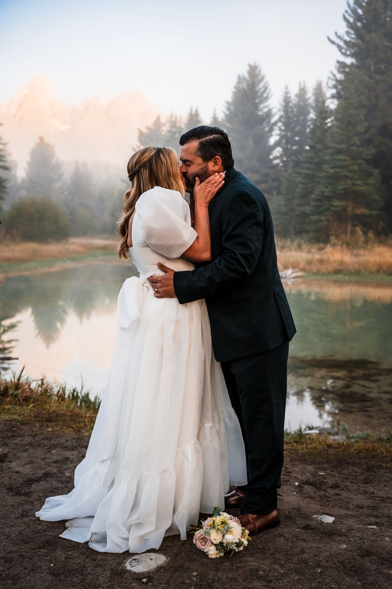 A romantic wedding couple shares a kiss by a misty lake at sunset with evergreen trees in the background.