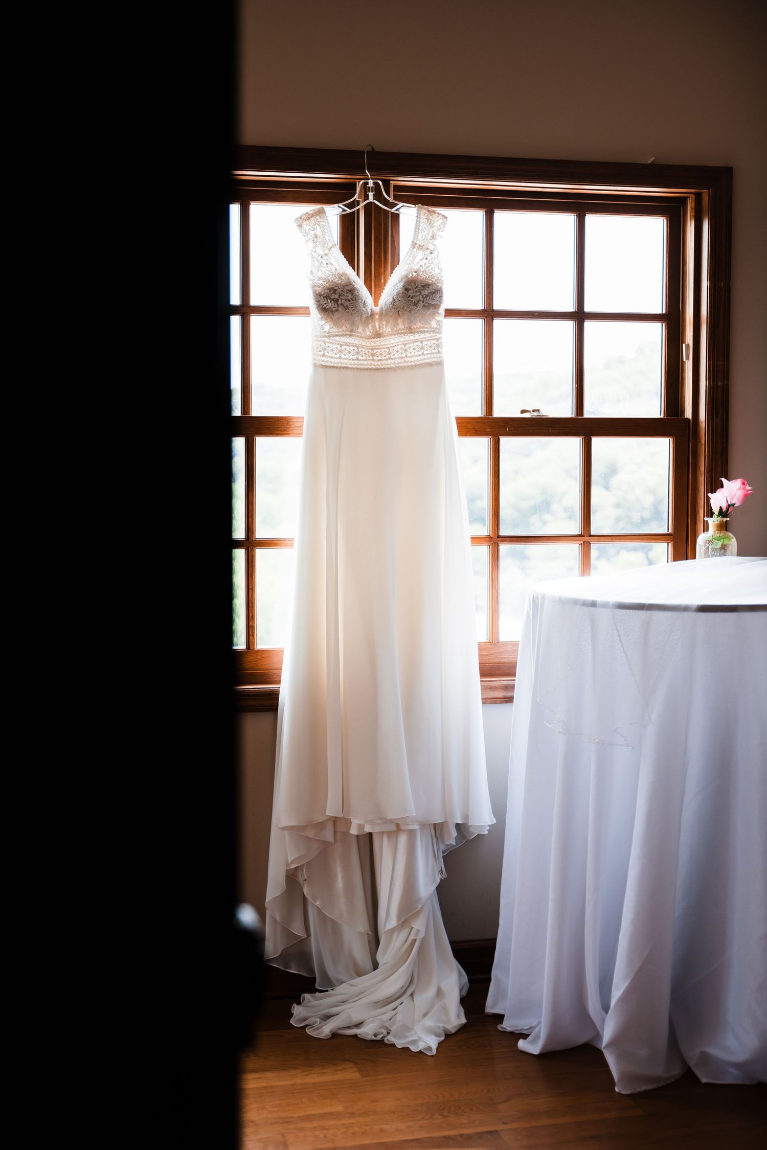 White wedding dress hanging by windows in a rustic indoor setting with natural light.