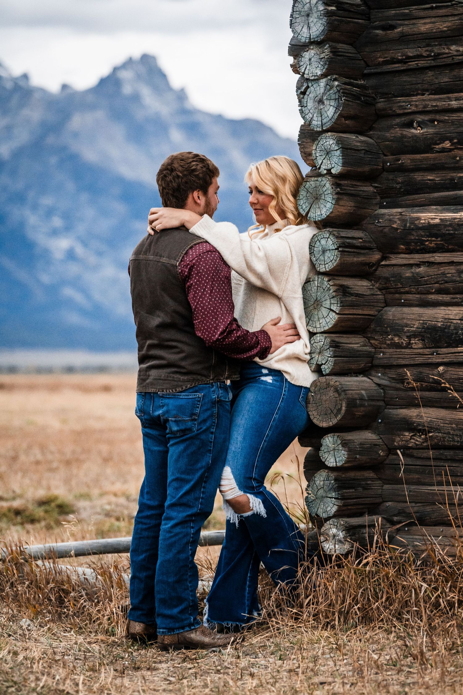 A couple shares a romantic moment against a rustic log cabin wall with mountains in the background.