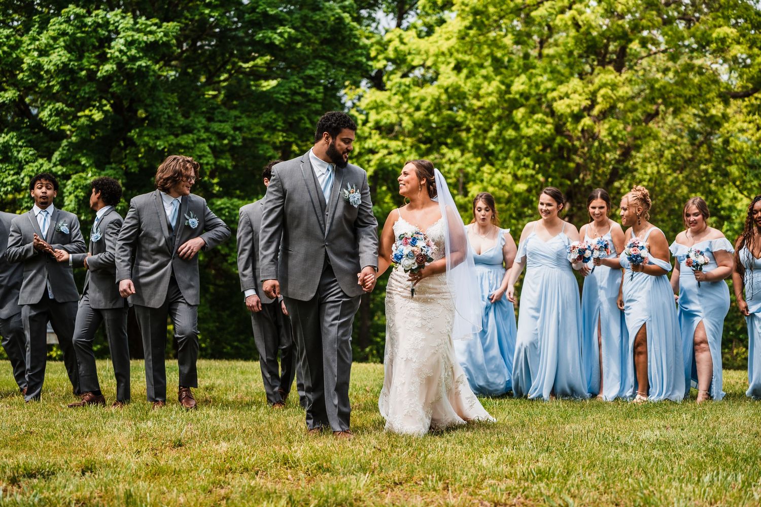 Wedding party stands on grass with bridesmaids in light blue dresses and groomsmen in gray suits.