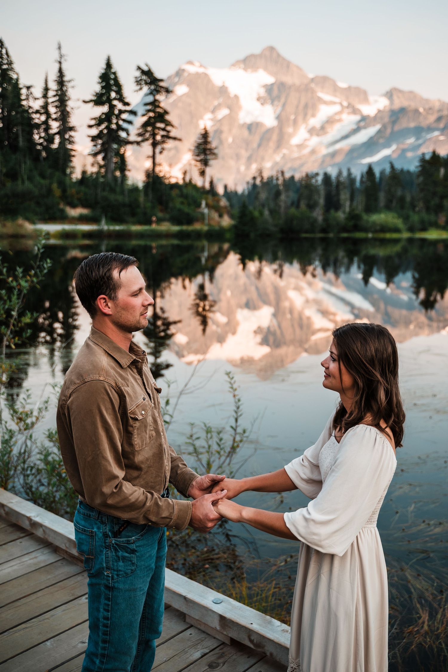 A romantic couple holds hands on a wooden deck overlooking a pristine mountain lake with snow-capped peaks at sunset.