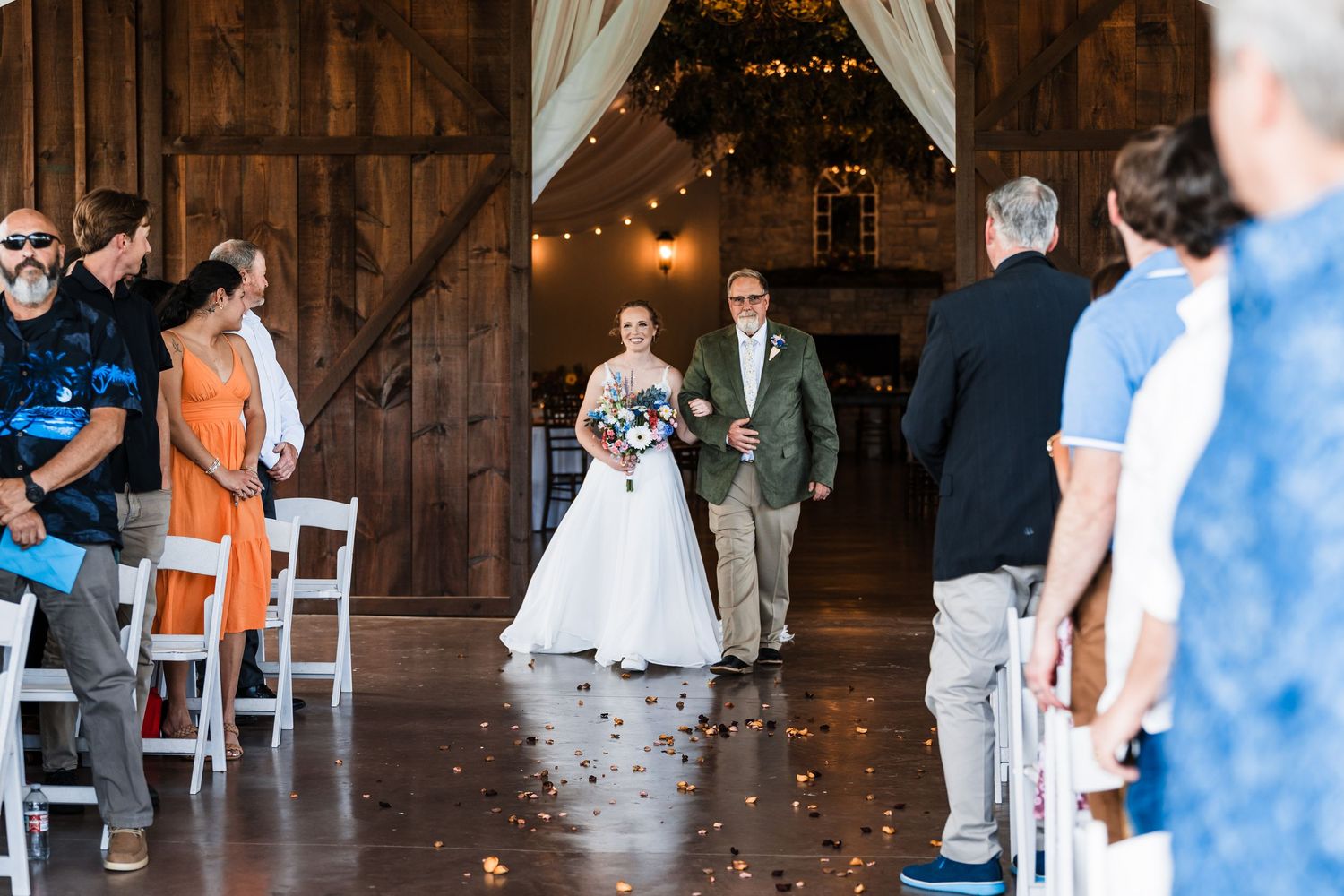 Bride walks down rustic barn wedding aisle with escort amid string lights and seated guests.