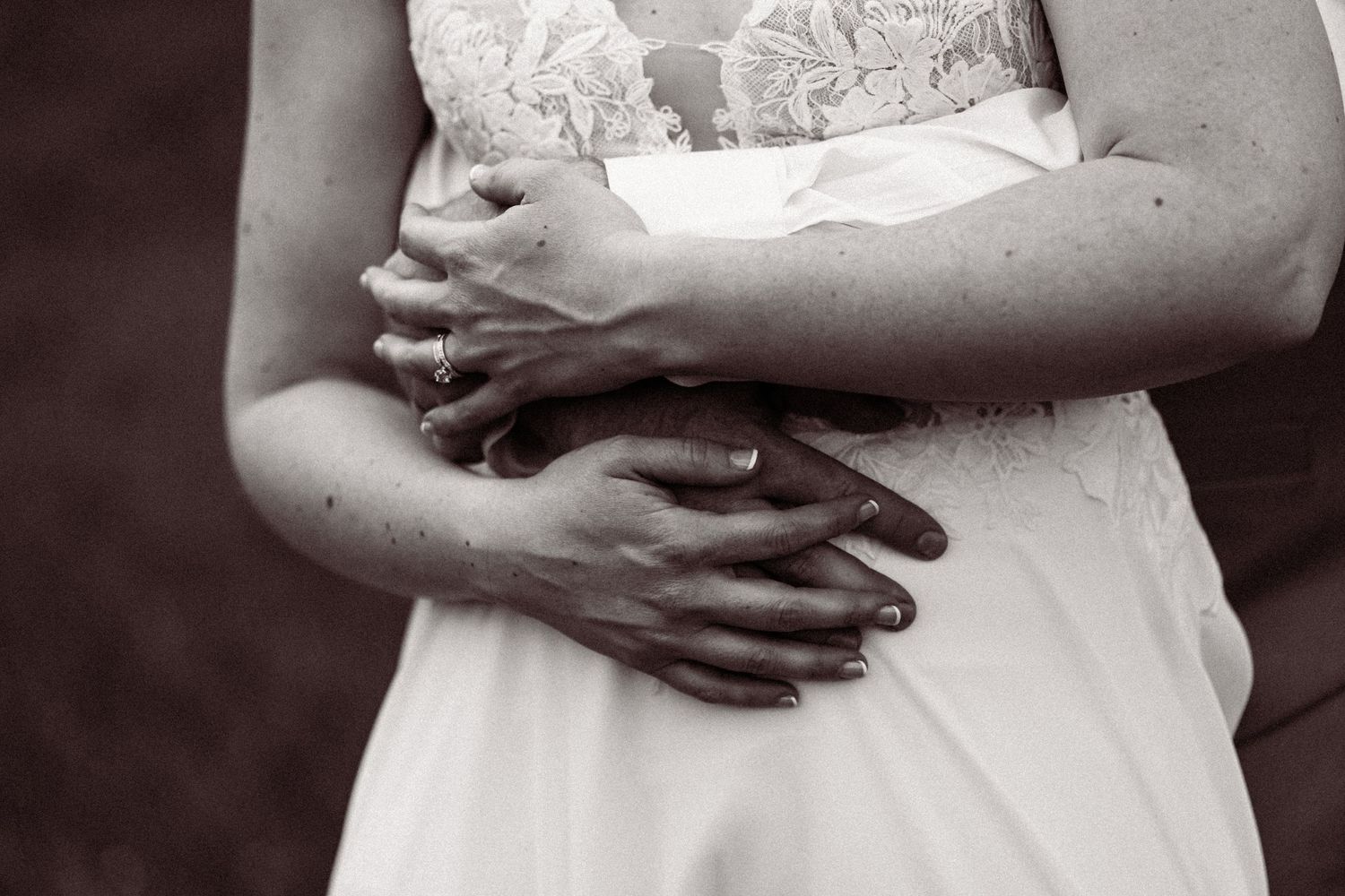 Black and white close-up of groom embracing bride in lace wedding dress, showing wedding rings.