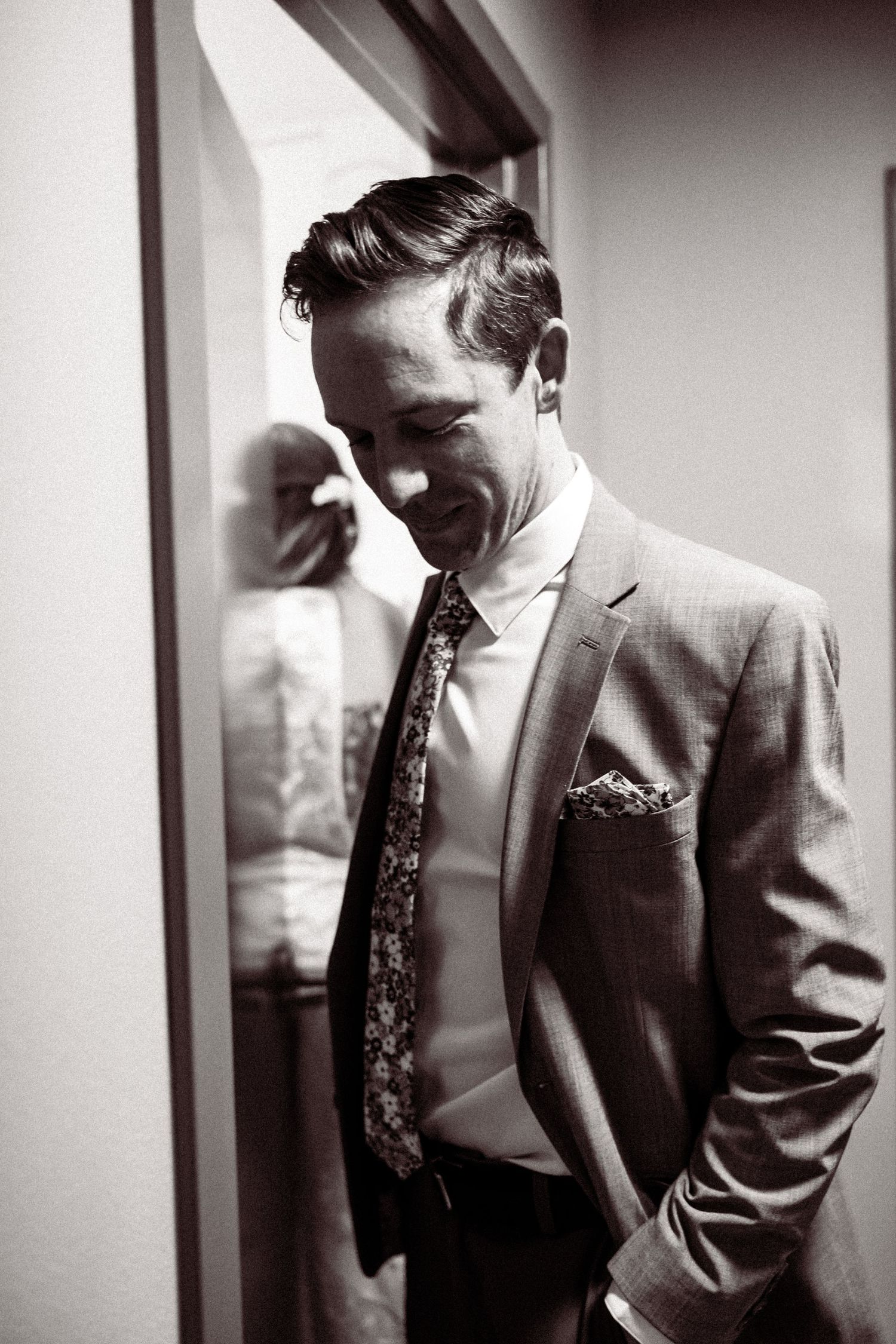 Dapper groom in gray suit and floral tie smiling in black and white wedding portrait.