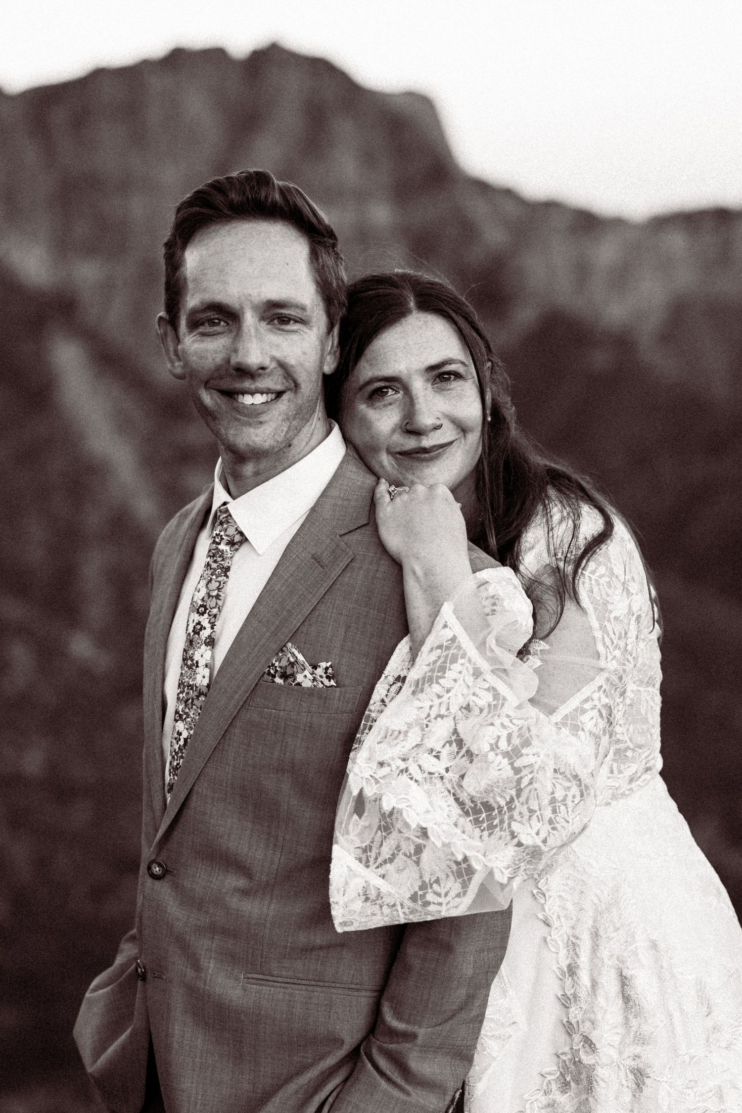 Black and white portrait of smiling groom in suit with bride leaning on his shoulder against mountain backdrop.