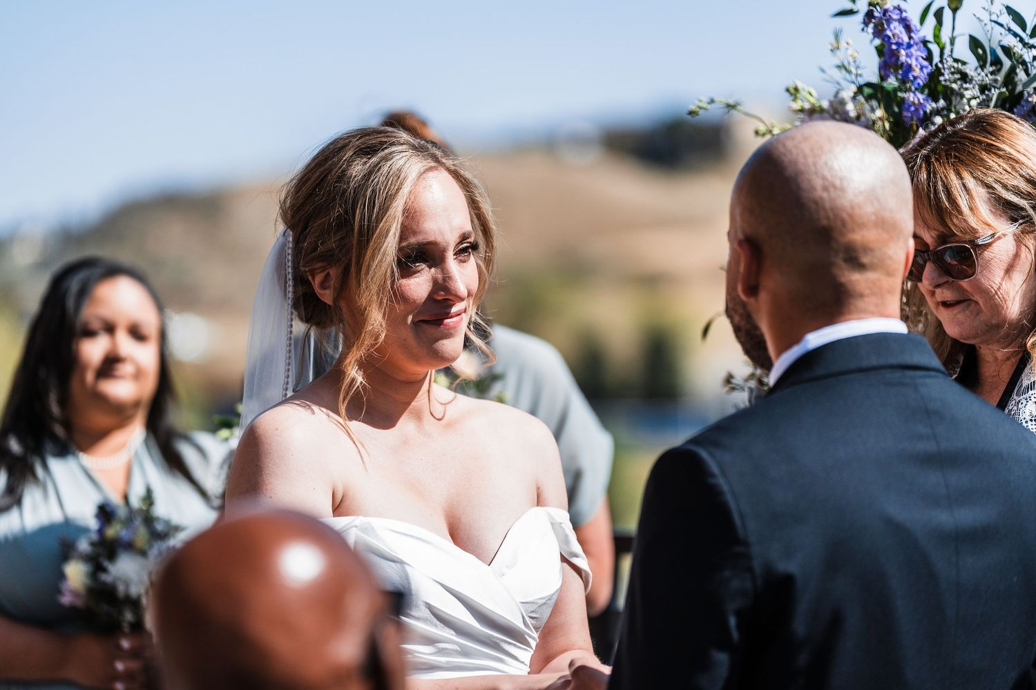 Smiling bride in white gown exchanges vows with groom at outdoor mountain wedding ceremony.