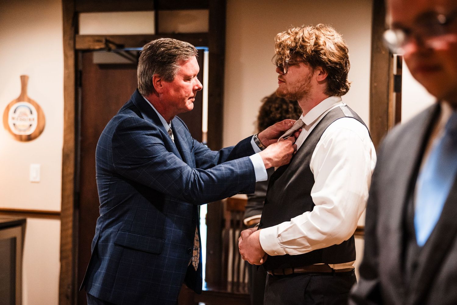 Older man in blue suit adjusting the tie of a young groom in vest at wedding preparation.
