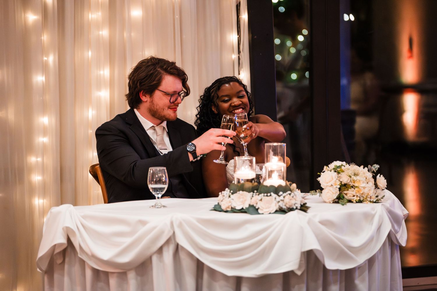 Newlyweds laugh and toast champagne at sweetheart table adorned with white florals and fairy lights.
