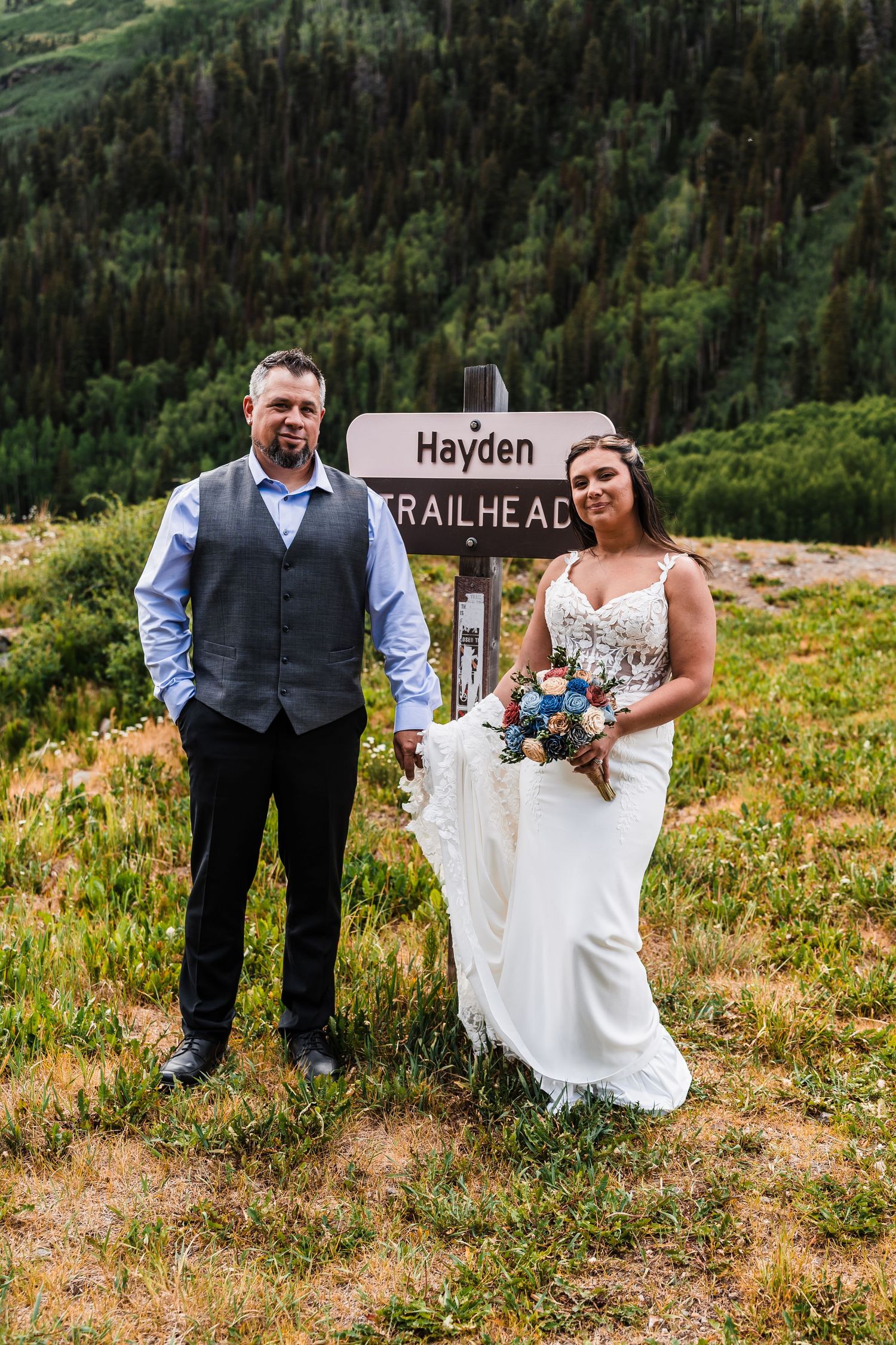 Bride and groom pose beside Hayden Trailhead sign in mountain meadow during outdoor elopement.