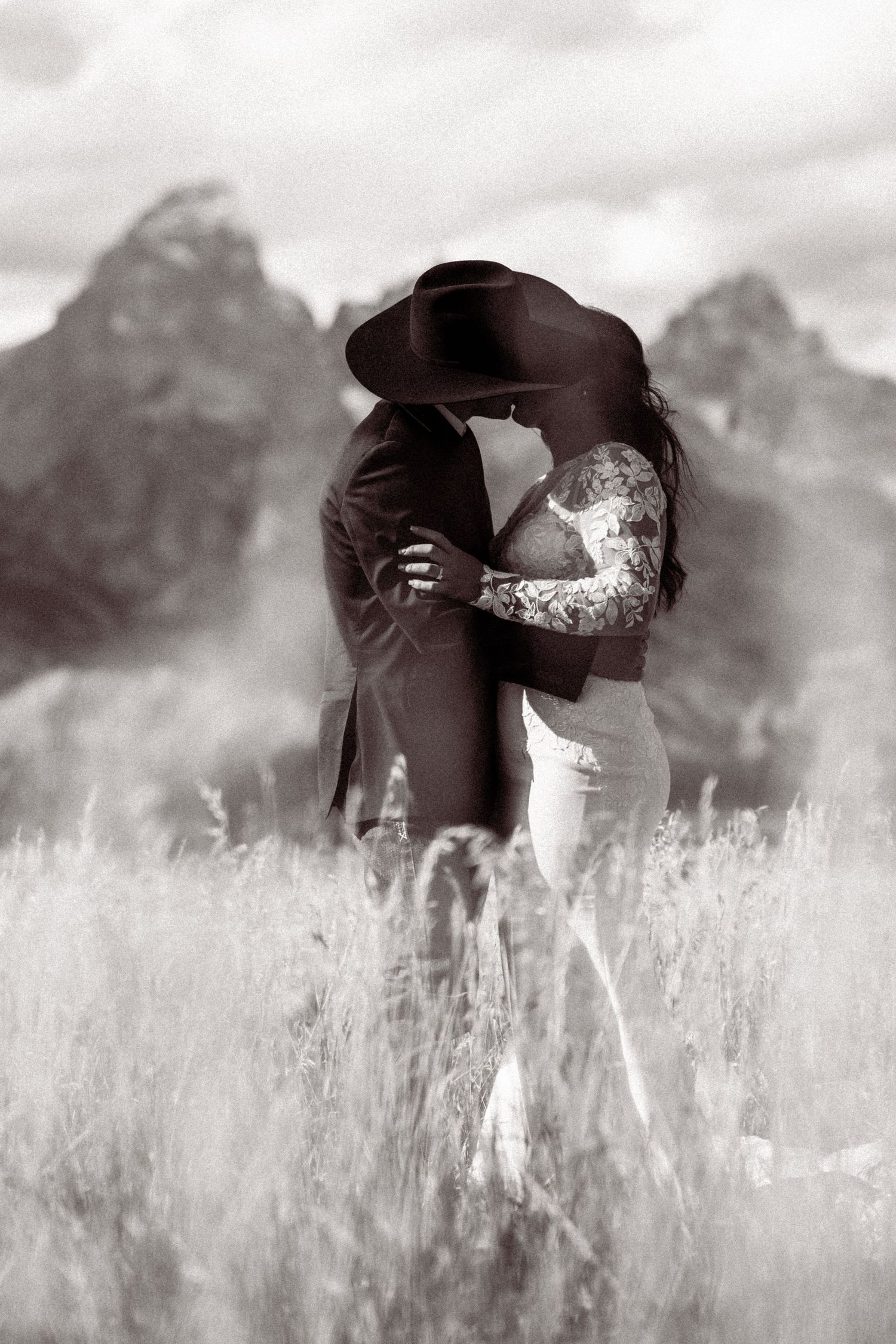 Black and white artistic photo of a couple embracing in a field with mountains in the background.