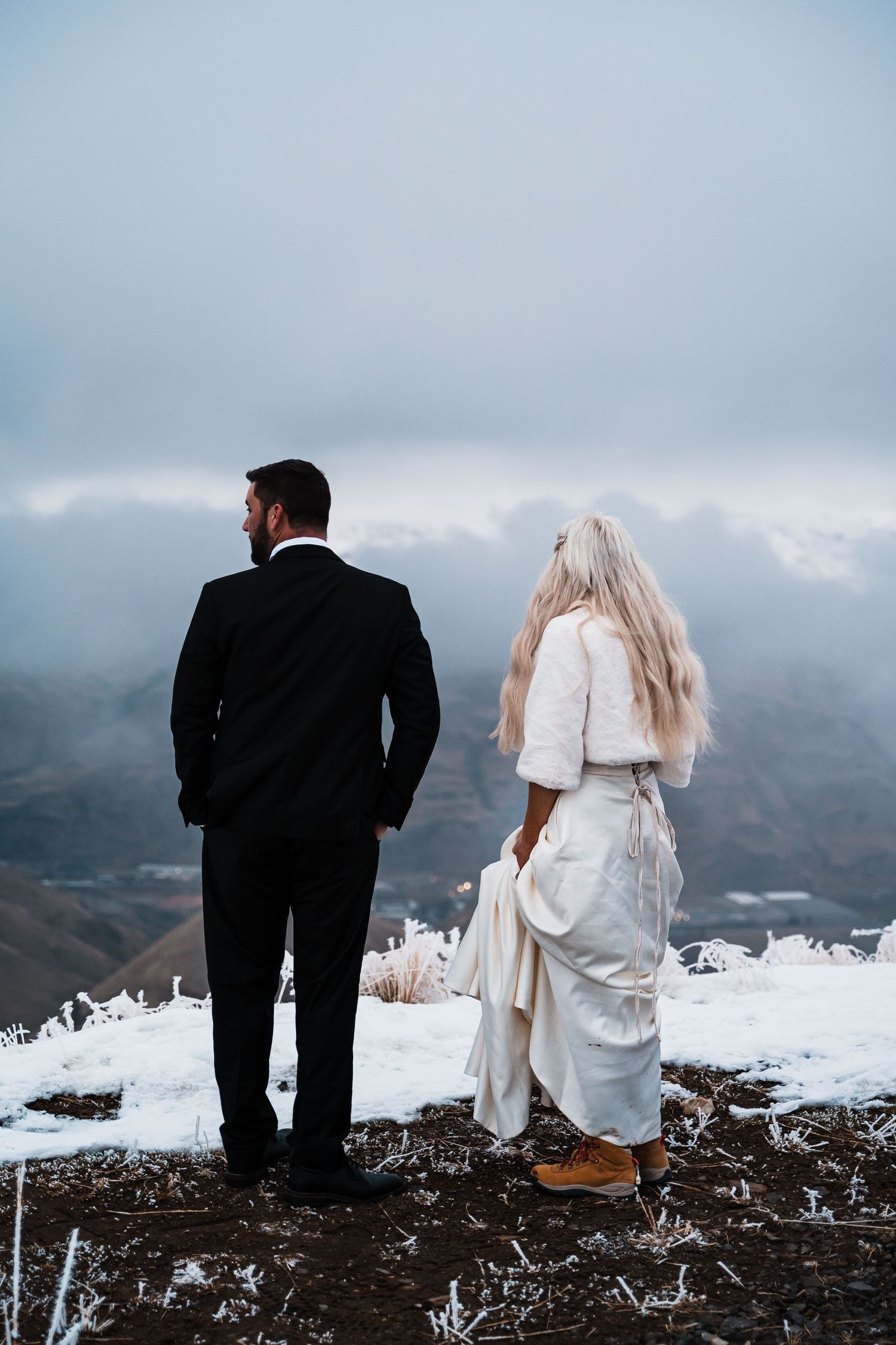 A couple in wedding attire sits together on a snowy mountain peak overlooking a dramatic cloudy landscape.