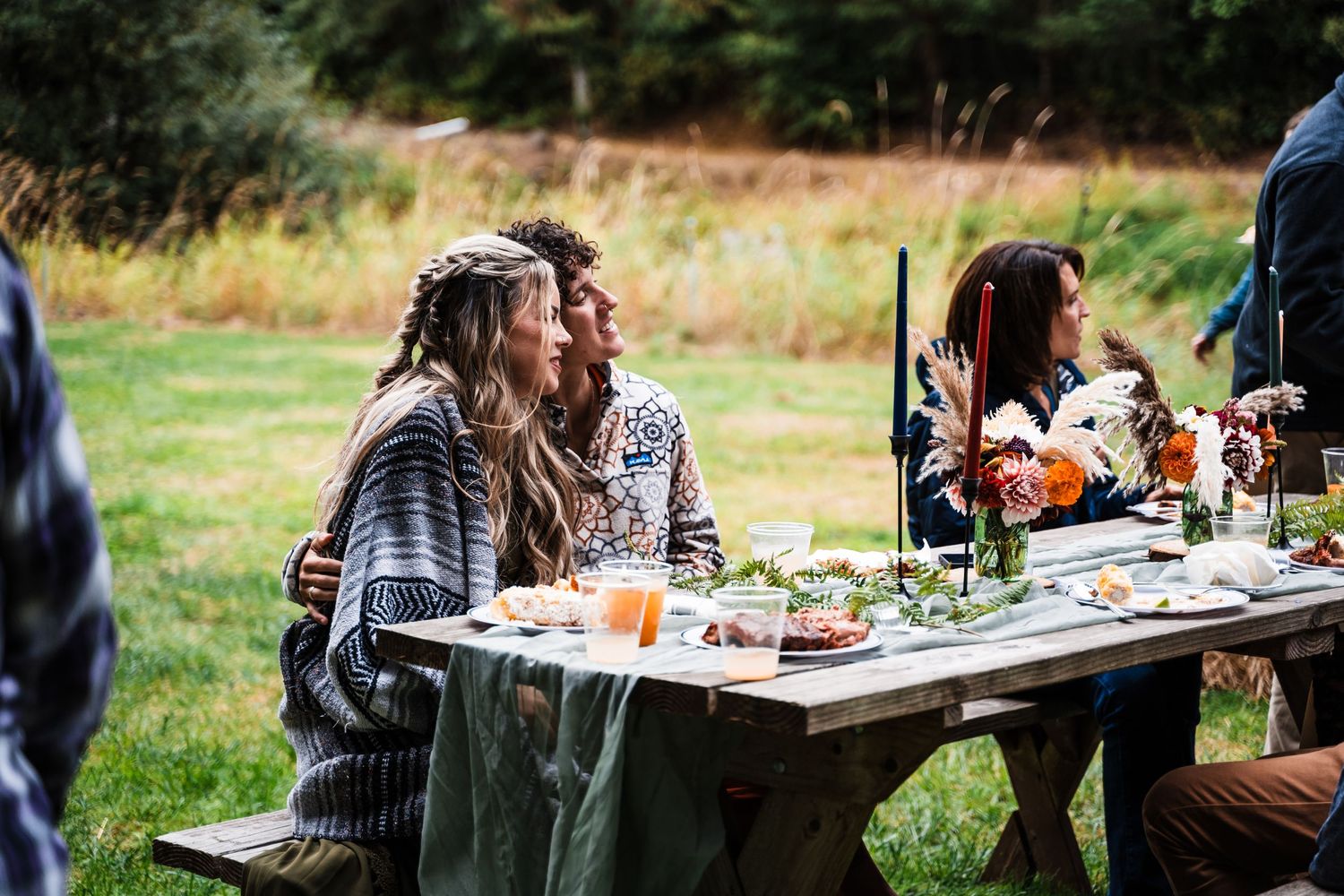 A casual outdoor gathering with people seated at a table enjoying food and drinks in nature.