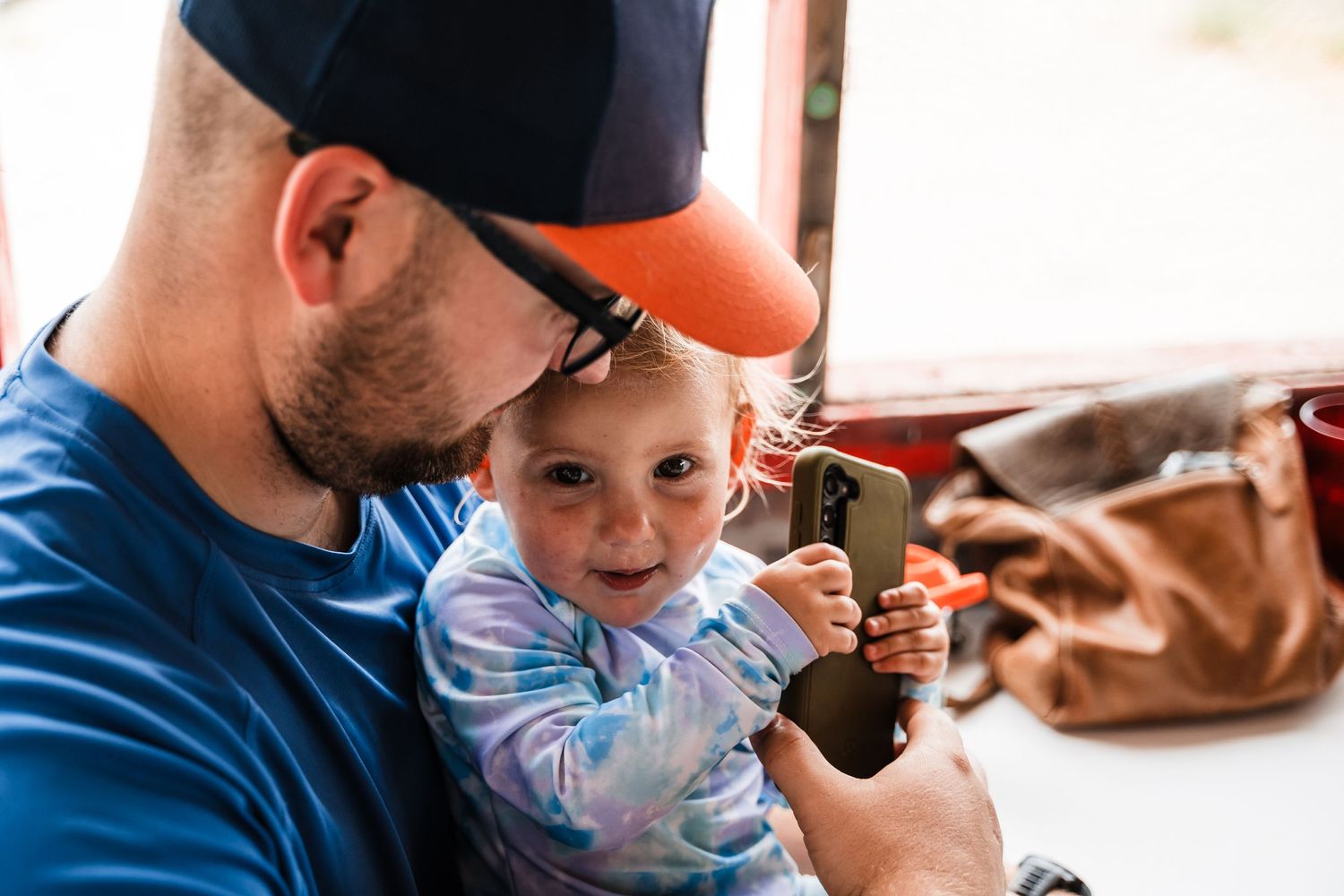 A young child looks at something being held while sitting with an adult wearing a baseball cap.