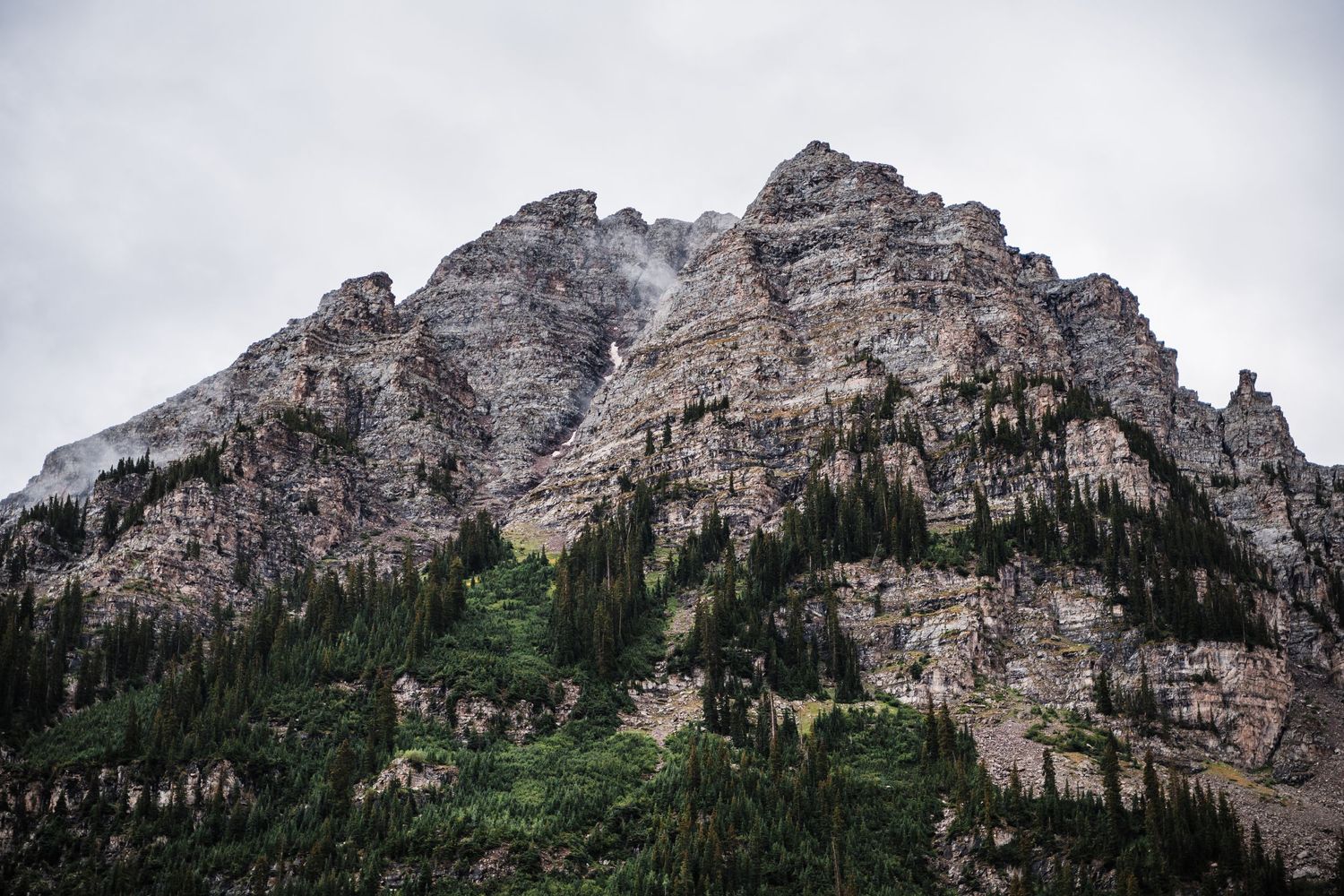 A majestic rocky mountain peak rises above evergreen forest under cloudy skies in a wilderness landscape.