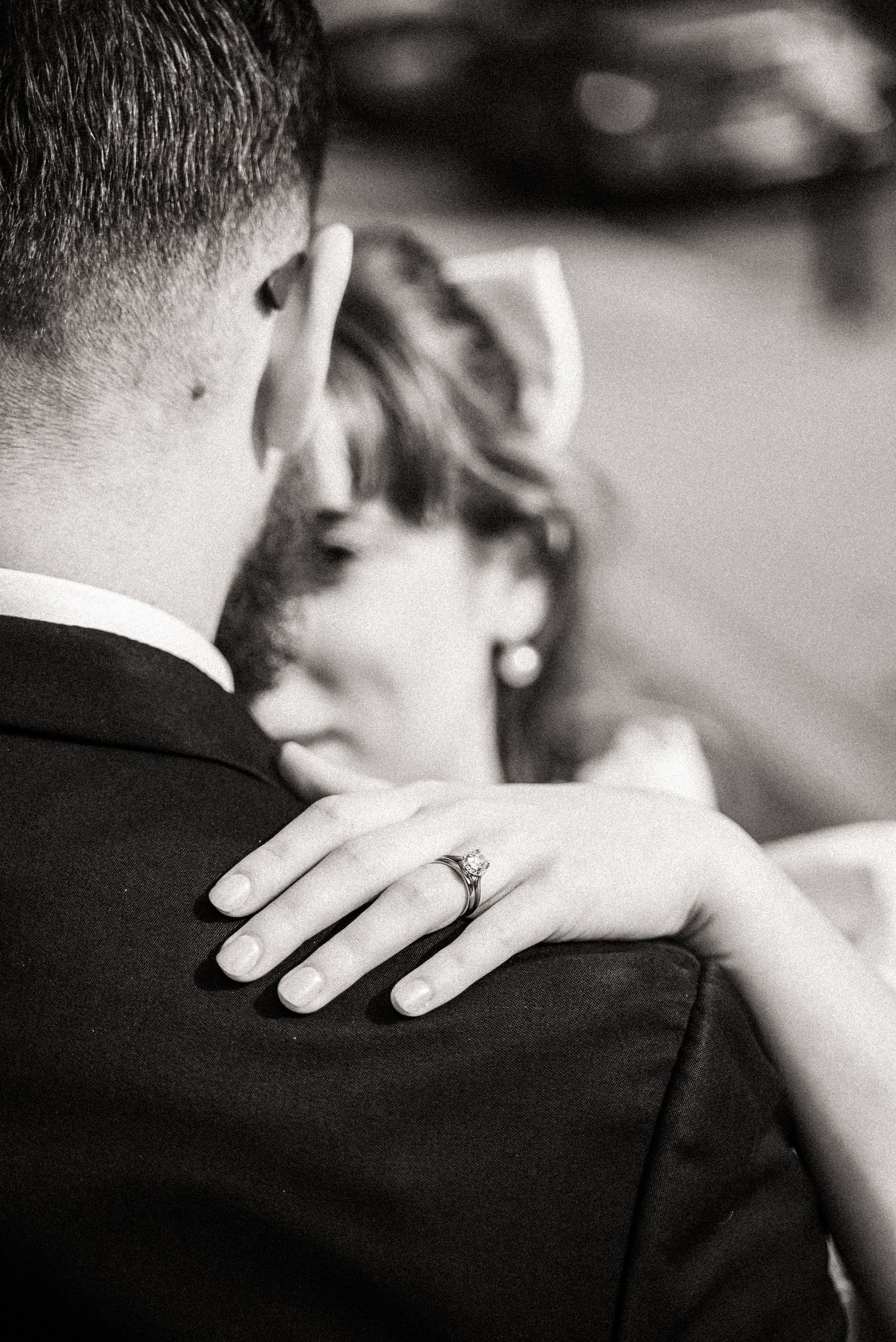 A black and white close-up of a couple embracing, showing a wedding ring on the hand resting on the groom's shoulder.