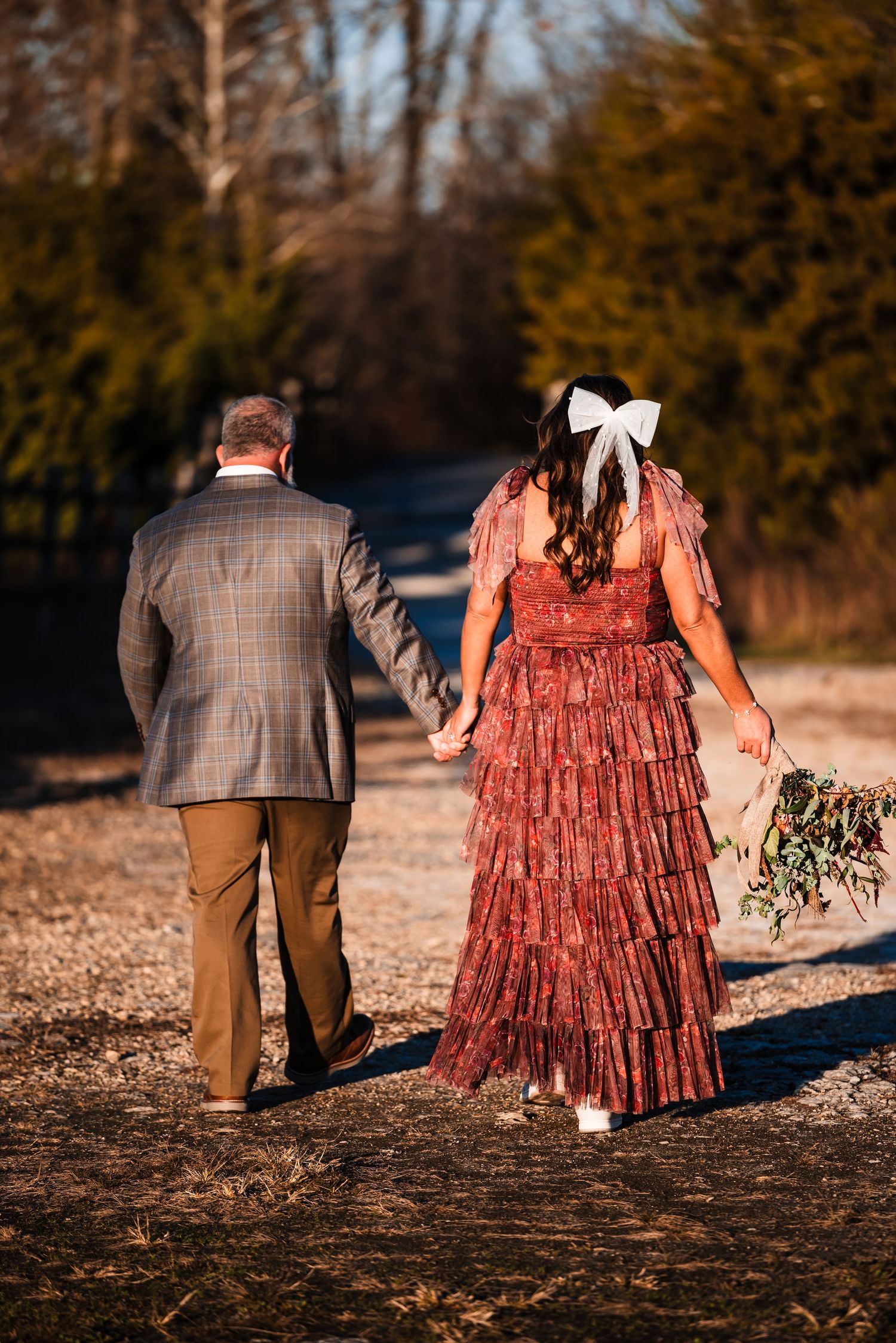 Couple walking hand in hand down path, wearing plaid suit and burgundy dress.