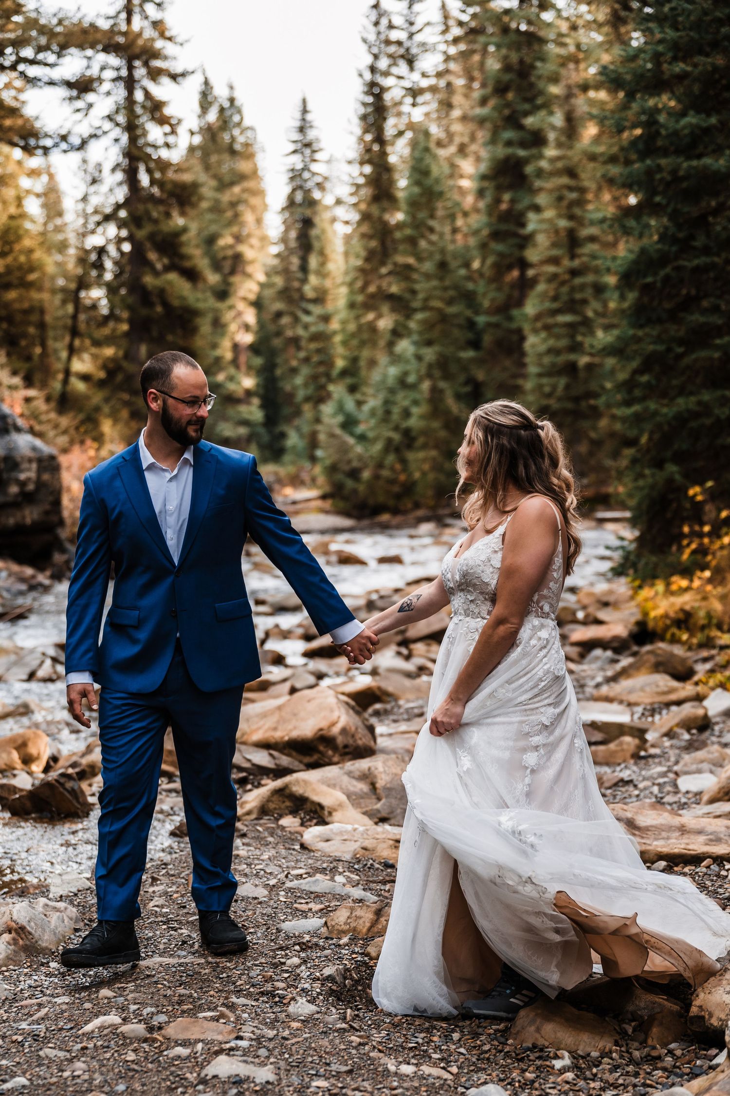 A couple traverses rocky terrain in formal wedding attire among pine trees.