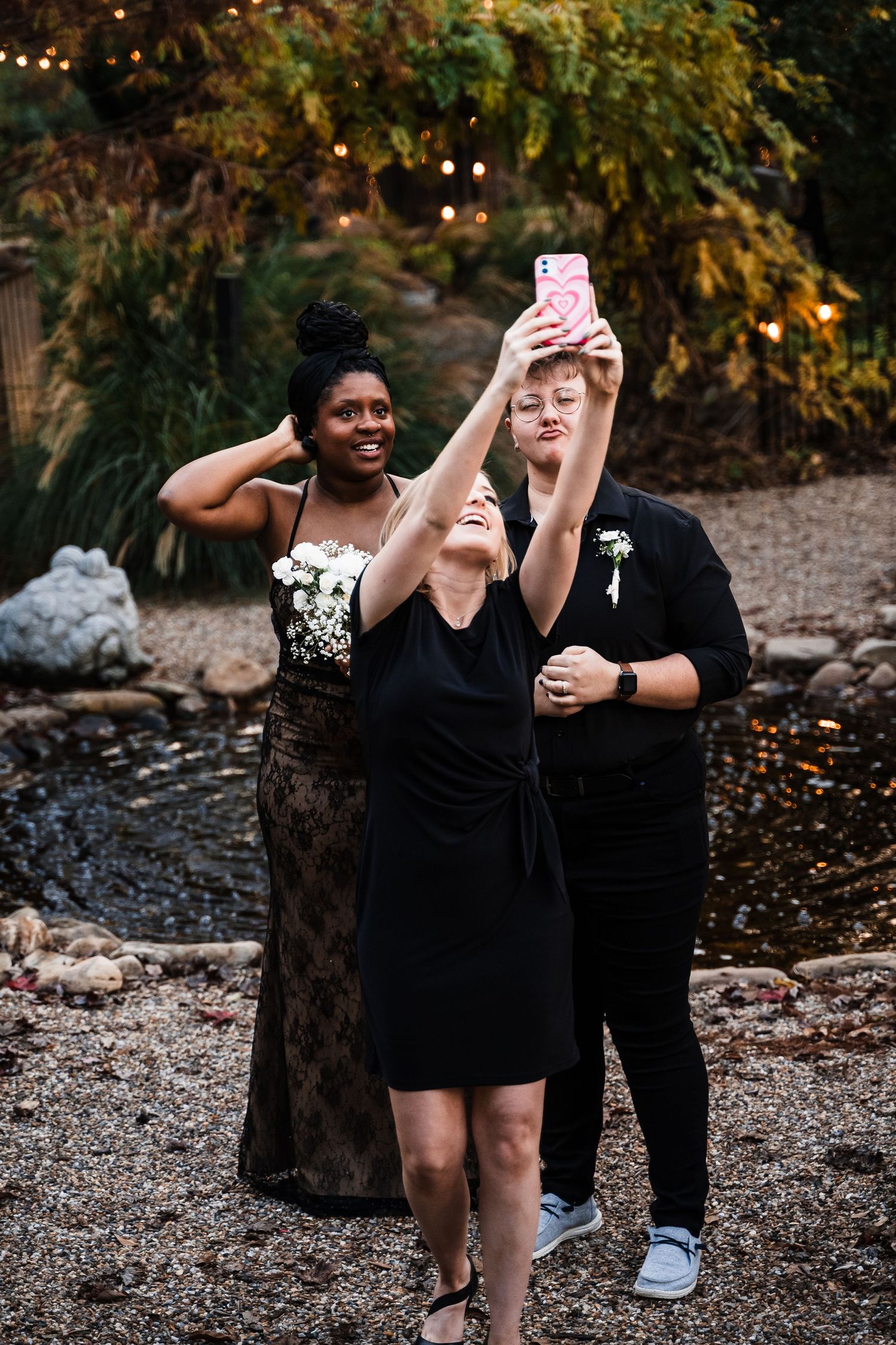 A group taking a selfie together at an outdoor wedding celebration with string lights and autumn foliage in the background.