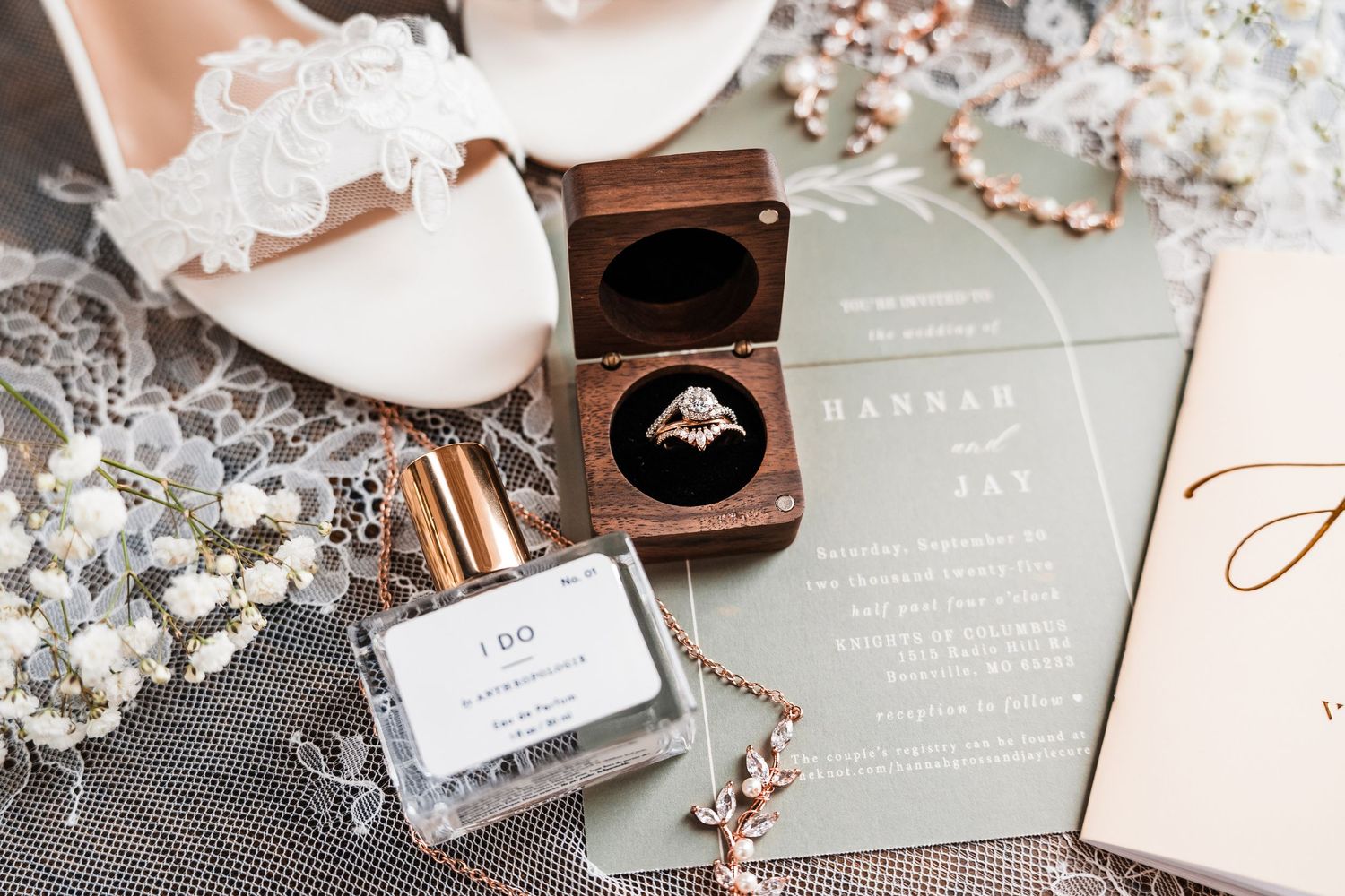Wedding invitation suite with sage green cards, white flowers, and rose gold ring box displayed on gray textured surface.