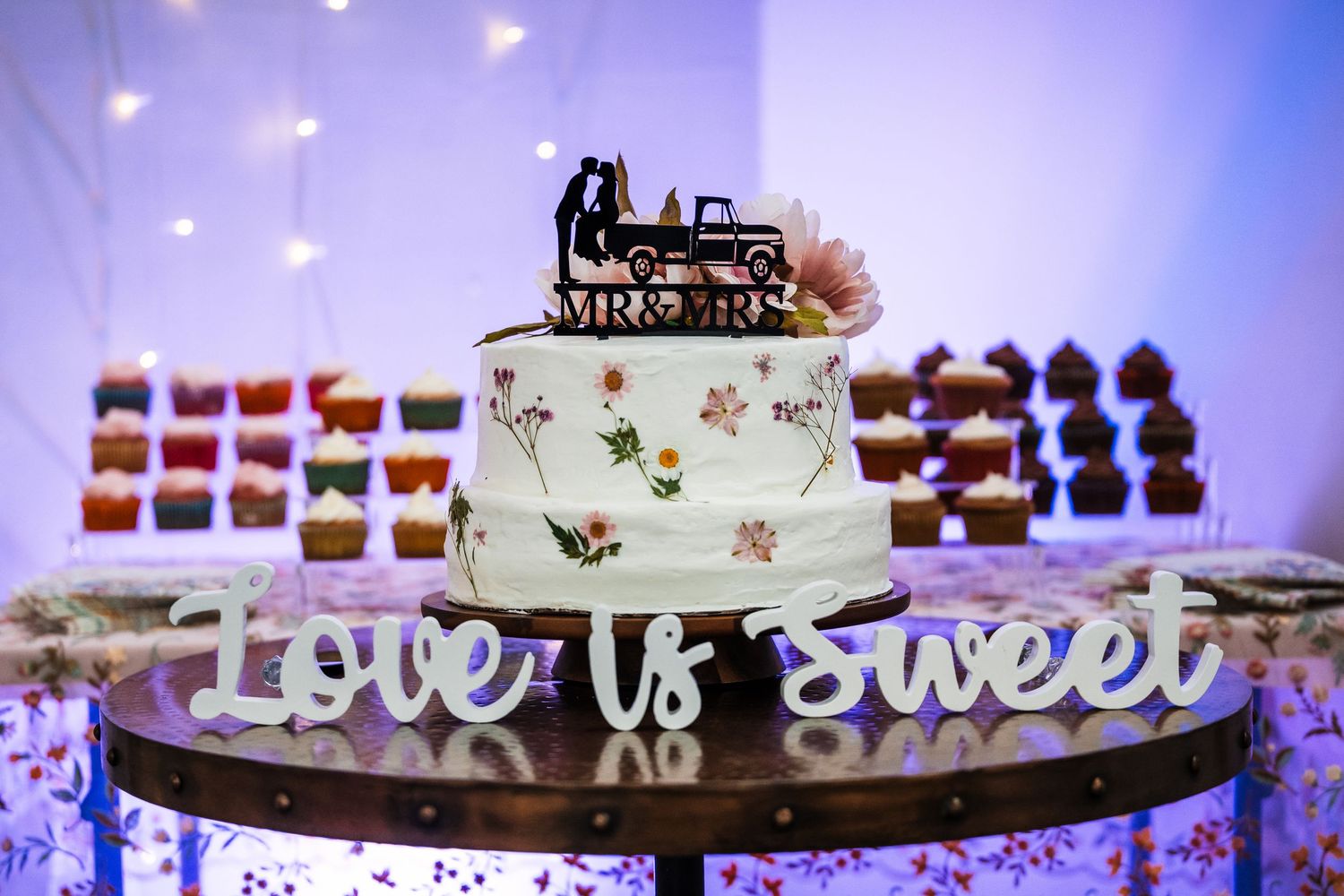White wedding cake with train topper and 'Love is Sweet' sign displayed on illuminated dessert table.