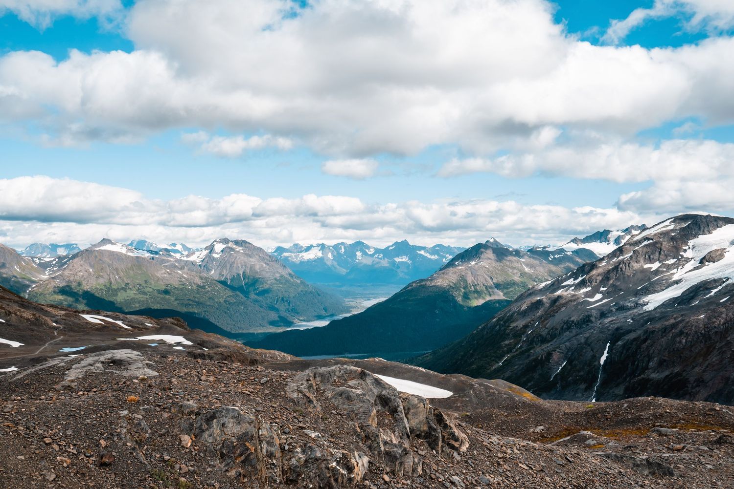Snow-capped mountain peaks and rugged terrain stretch across a dramatic alpine landscape under scattered clouds.