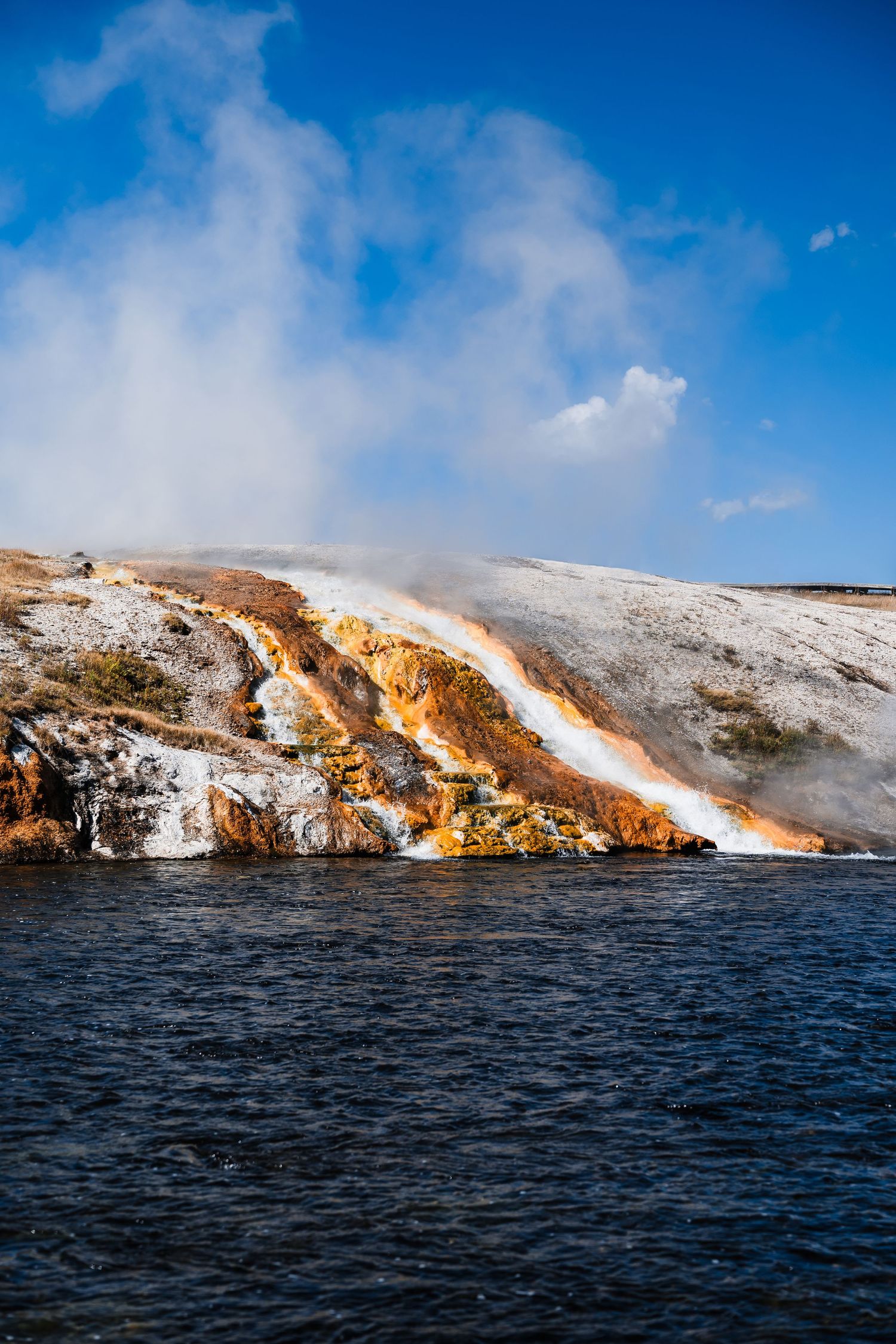 Colorful mineral deposits and steam flow down snowy hillside into dark waters at Yellowstone's thermal features.