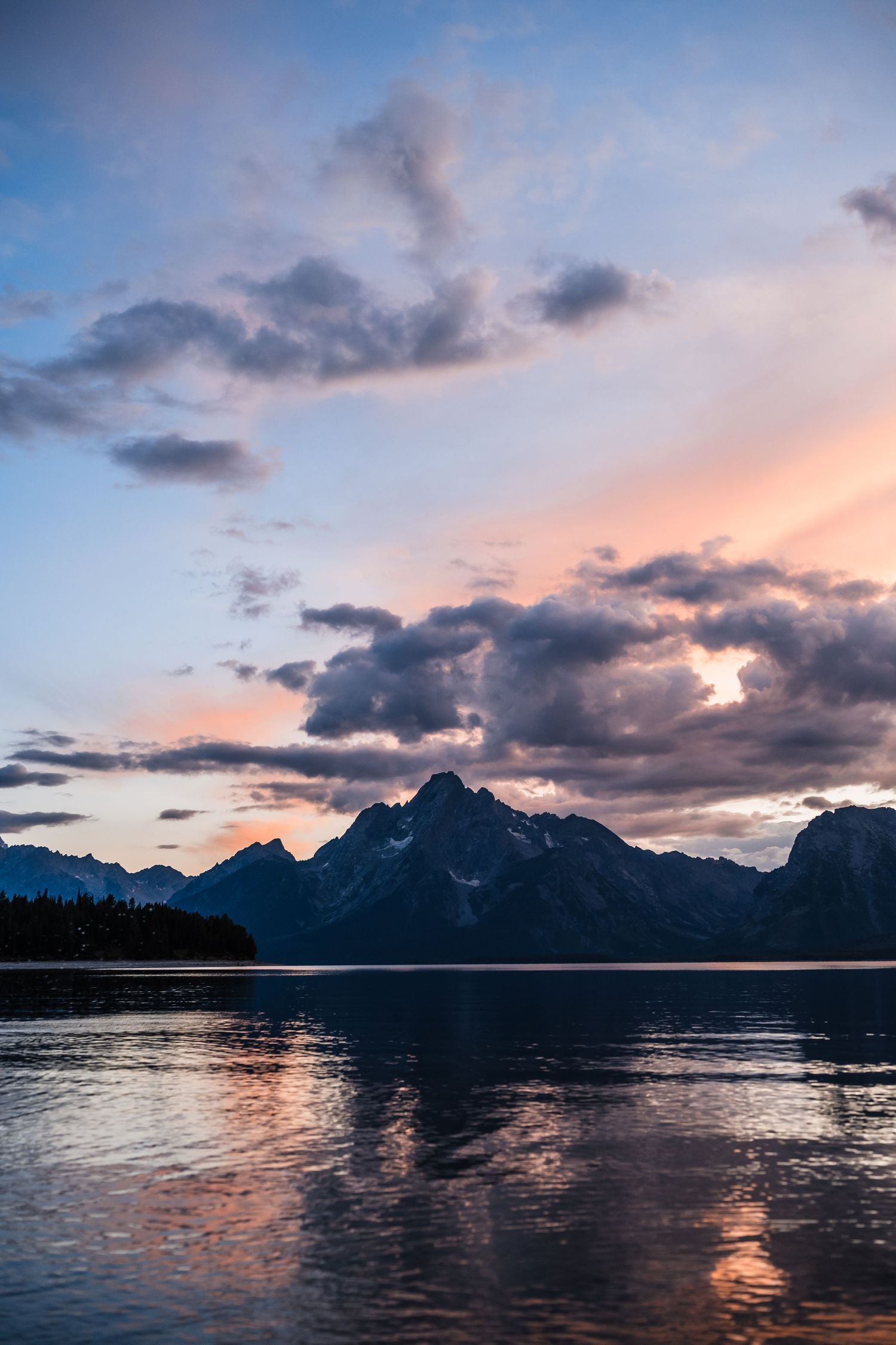 Dramatic sunset clouds reflect on calm lake waters with silhouetted mountain peaks in Grand Teton National Park.