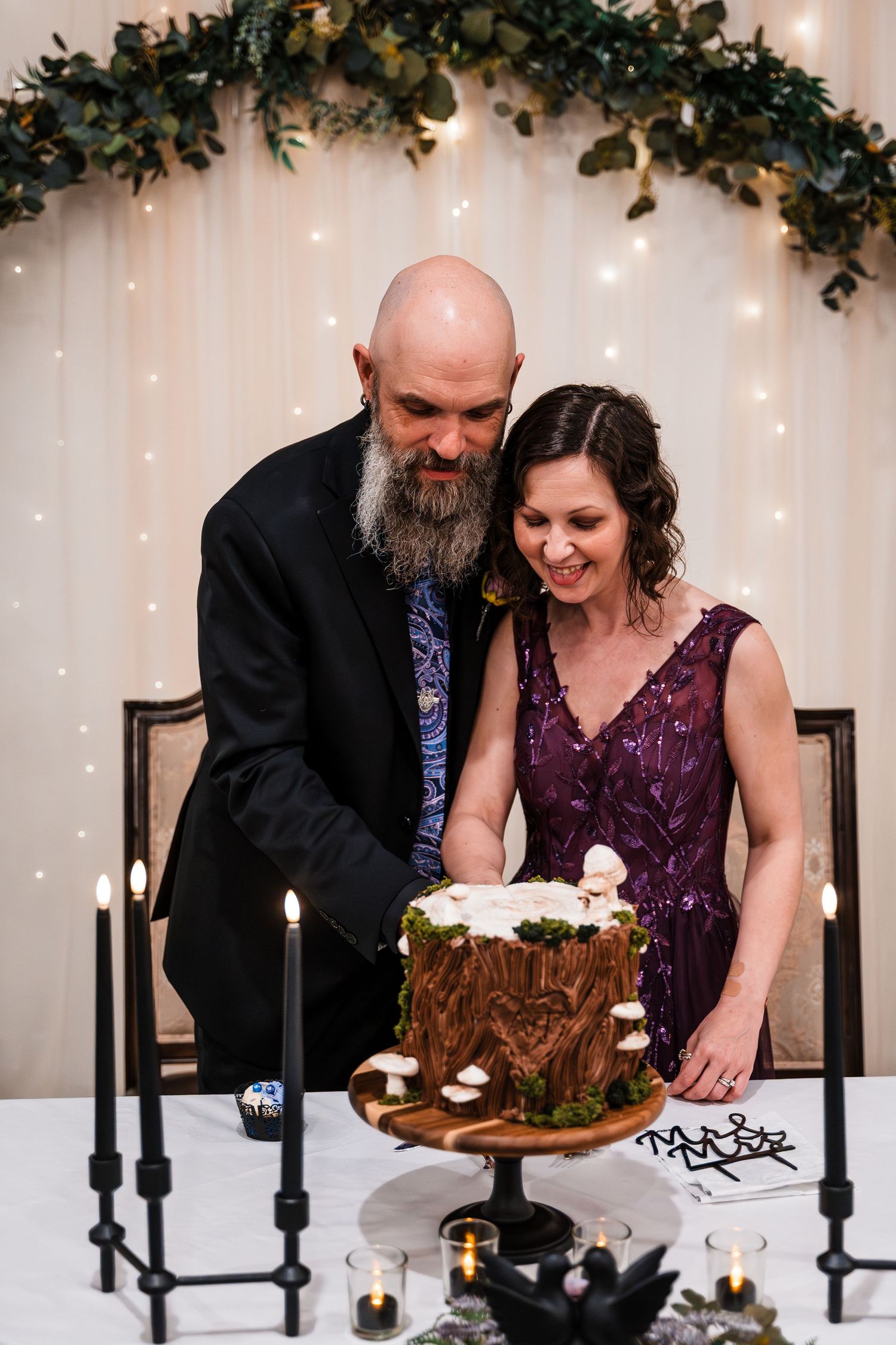 Bald bearded groom and brunette bride in purple dress cutting rustic woodland wedding cake with candles.