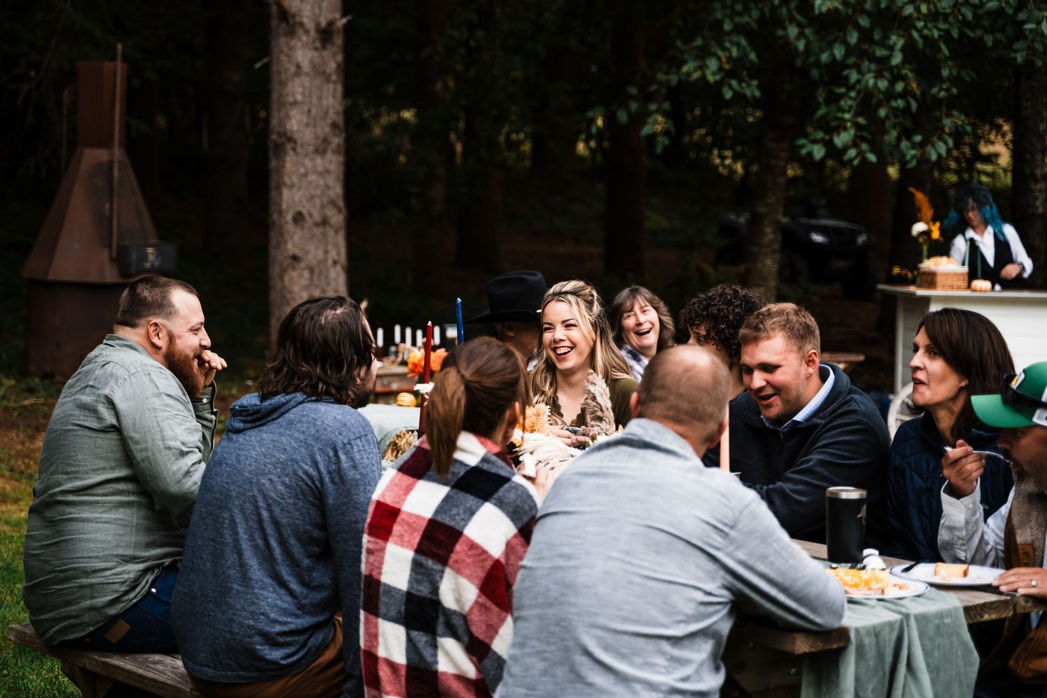 Friends gather at an outdoor table for drinks and conversation in a wooded setting.