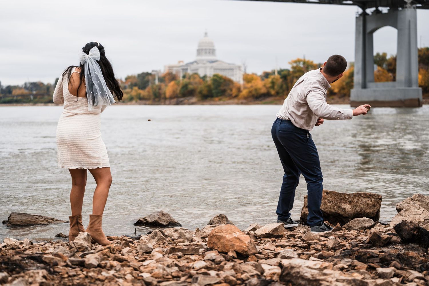 Two people searching through rocks on a riverbank with a city skyline in the background.