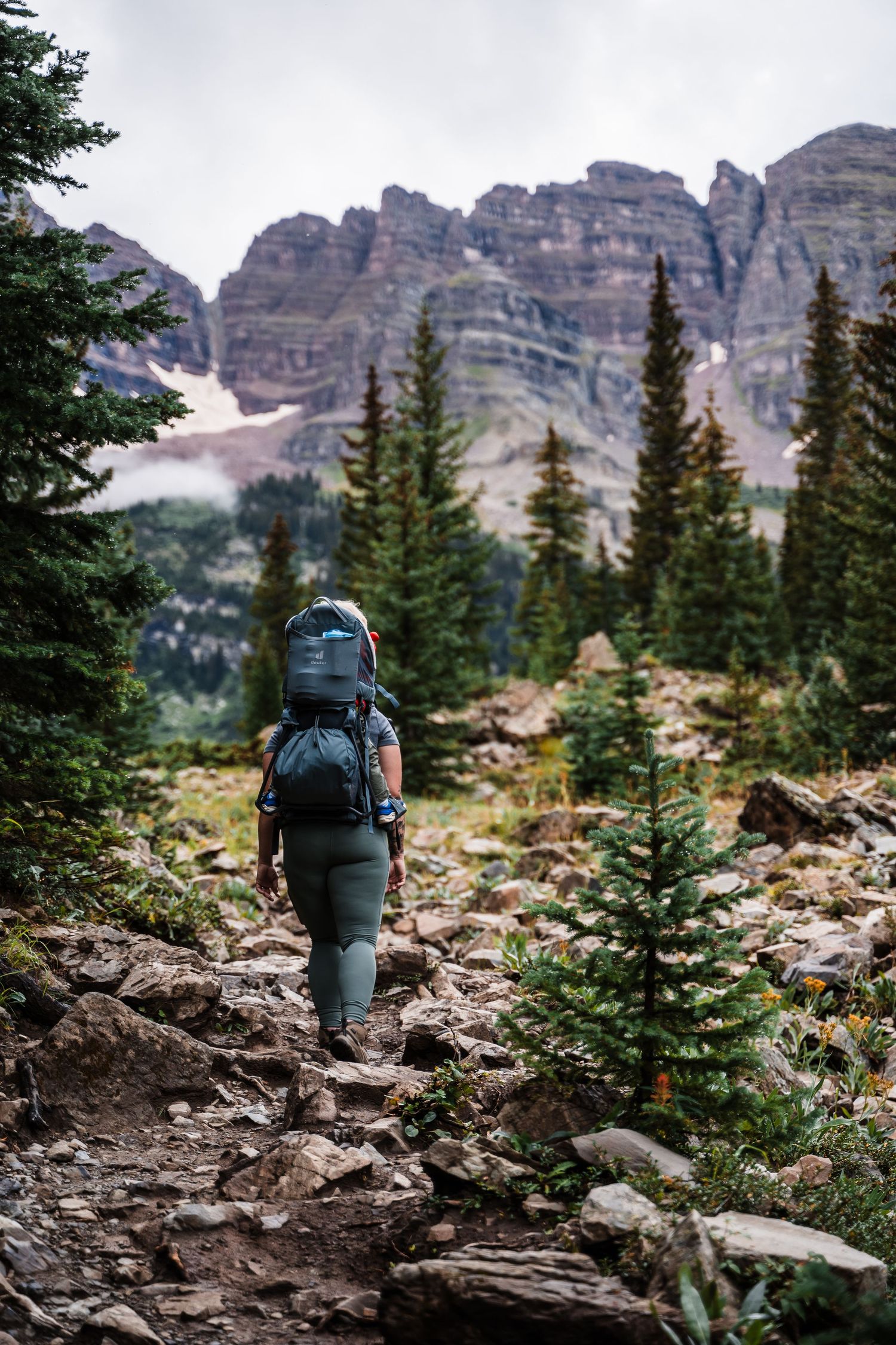 Hiker ascending rocky mountain trail with dramatic jagged peaks and evergreen forest in background.