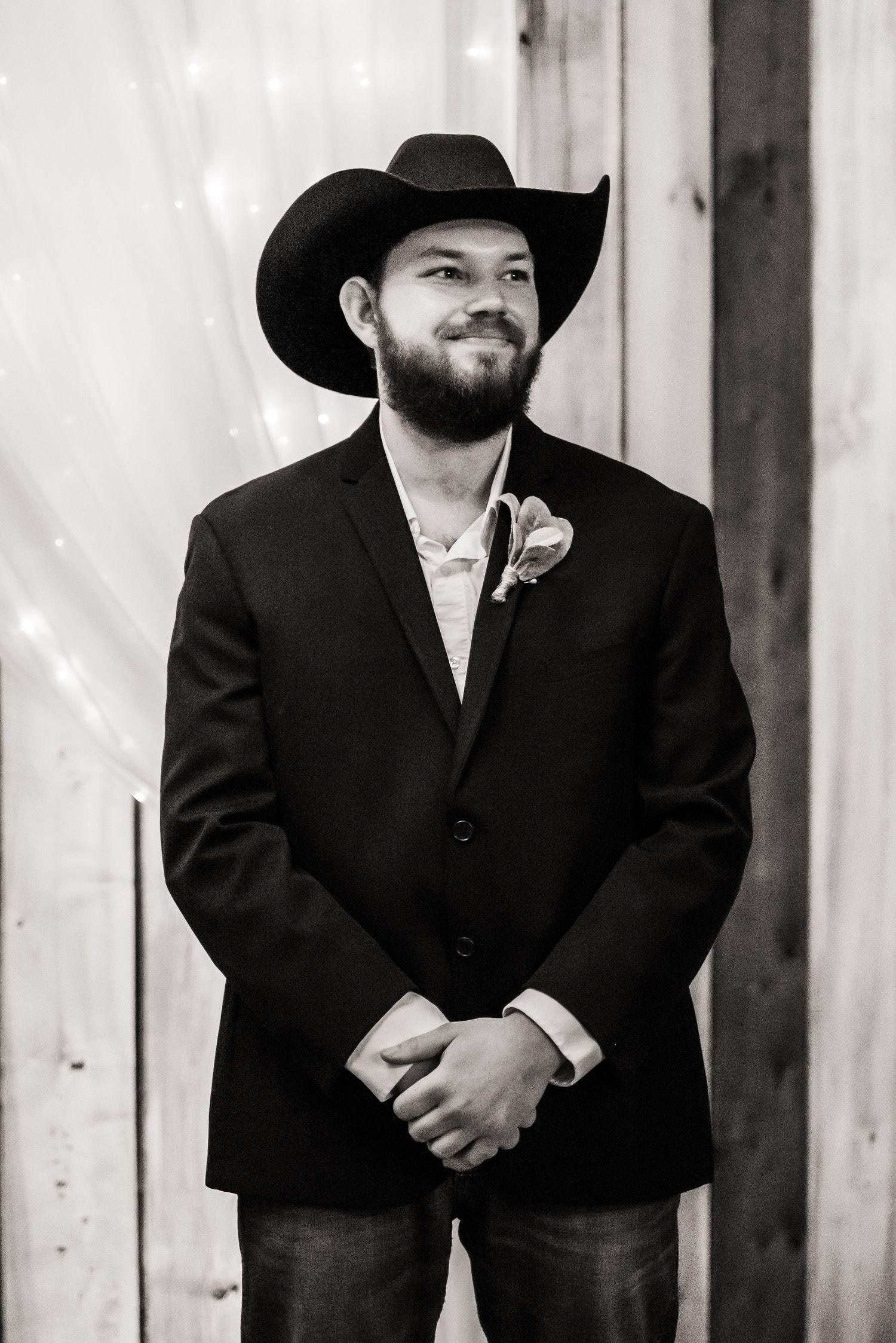 A formal portrait of a groom in a black suit jacket and cowboy hat with a boutonniere at a rustic wedding venue.