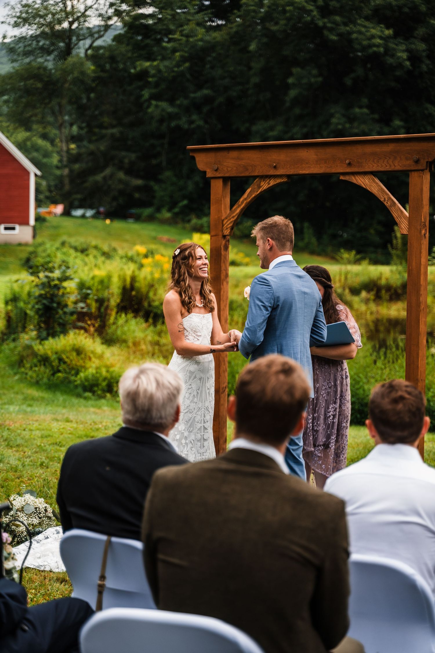 Intimate wedding ceremony under wooden arch with red barn visible in background.