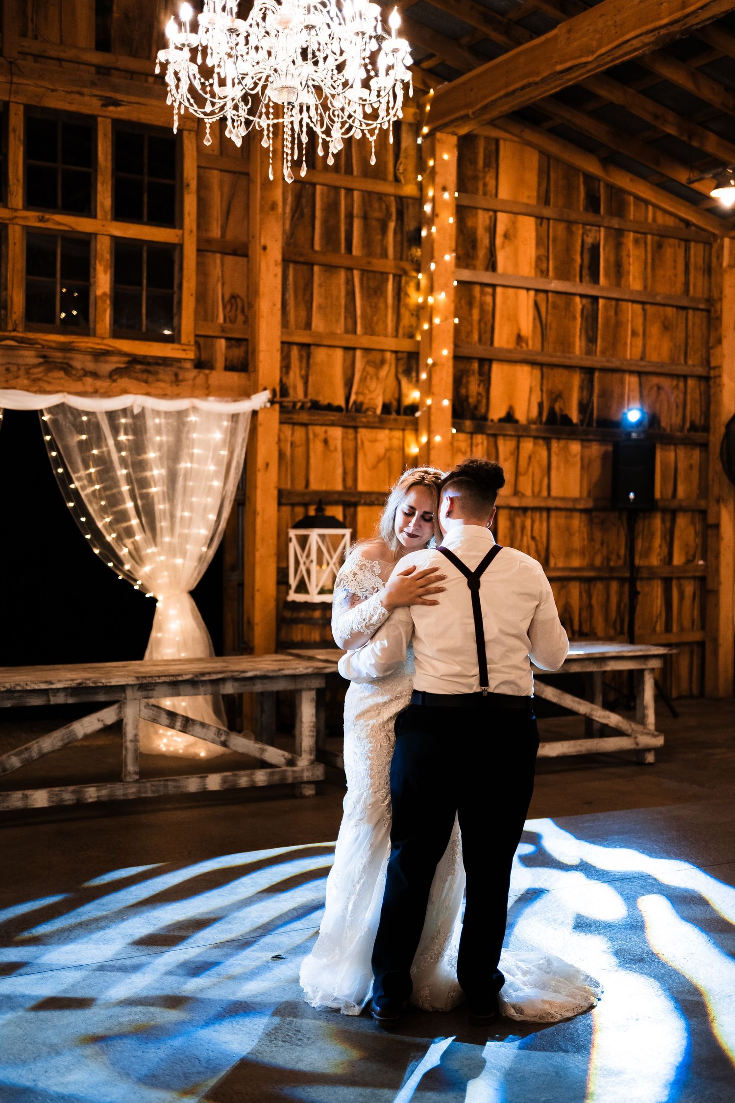 A couple dancing in a rustic barn wedding venue under a crystal chandelier with dramatic lighting on the floor.