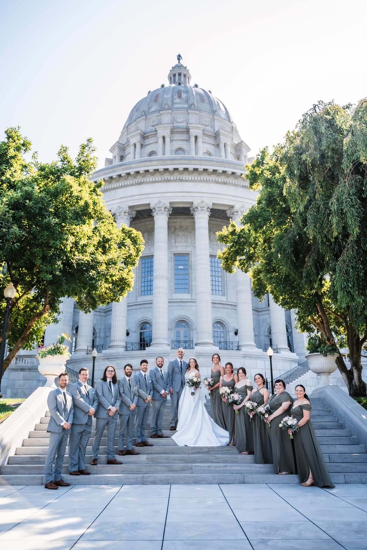 A wedding party poses on the steps of a grand domed government building with classical architecture.