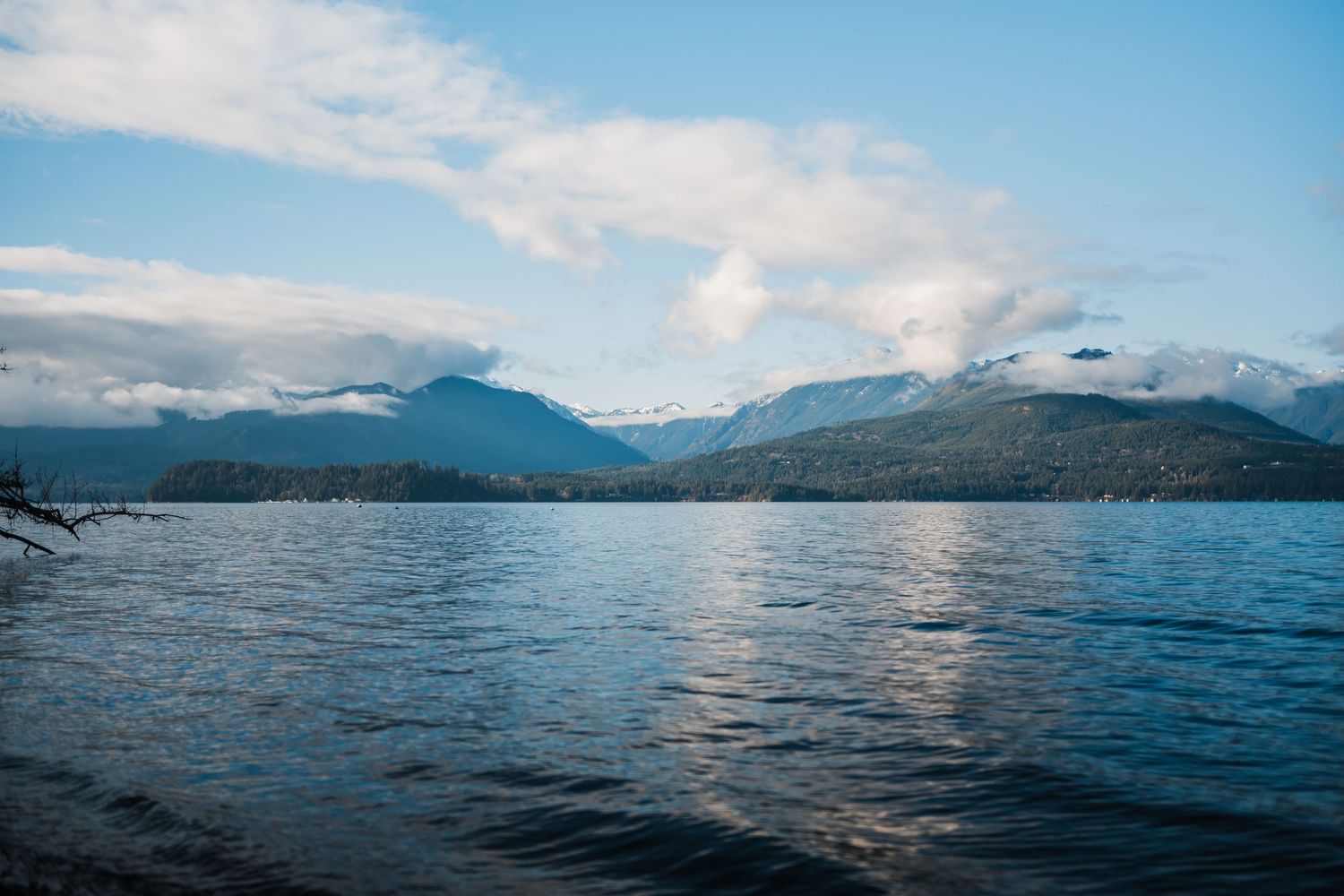 A serene mountain lake reflects blue skies and snow-capped peaks in the distance on a cloudy day.