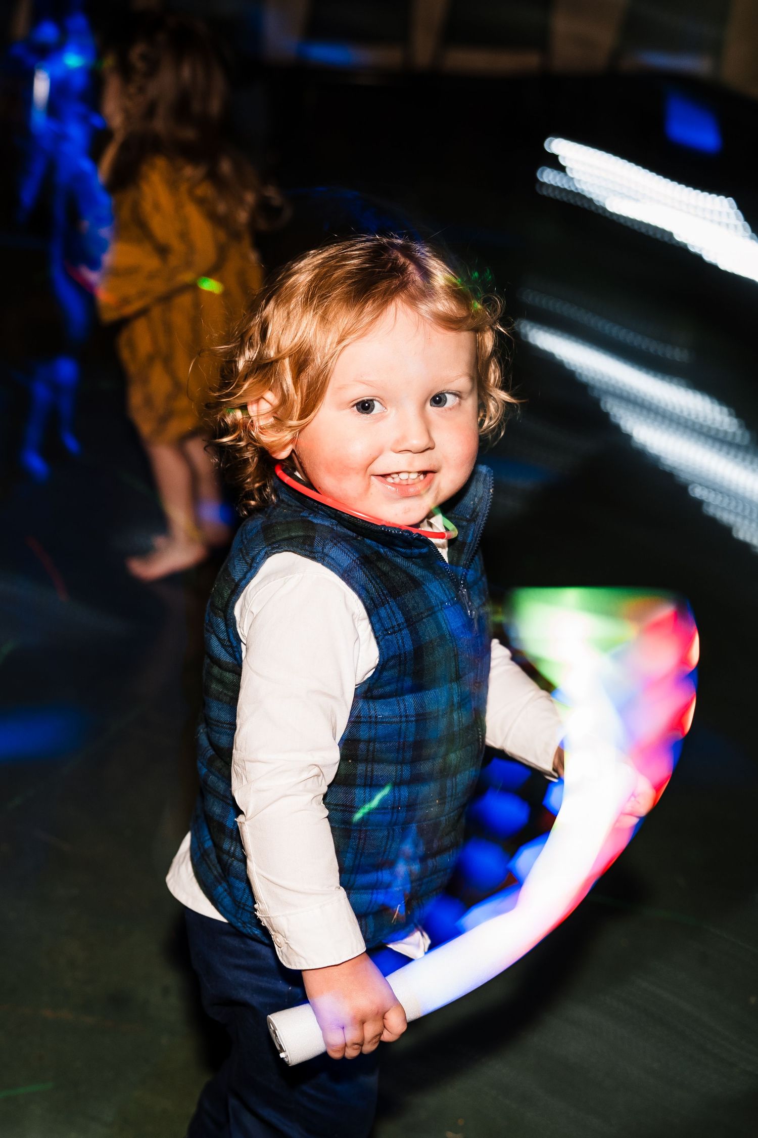 A small child in a blue vest plays with colorful light-up toys at a dark reception.