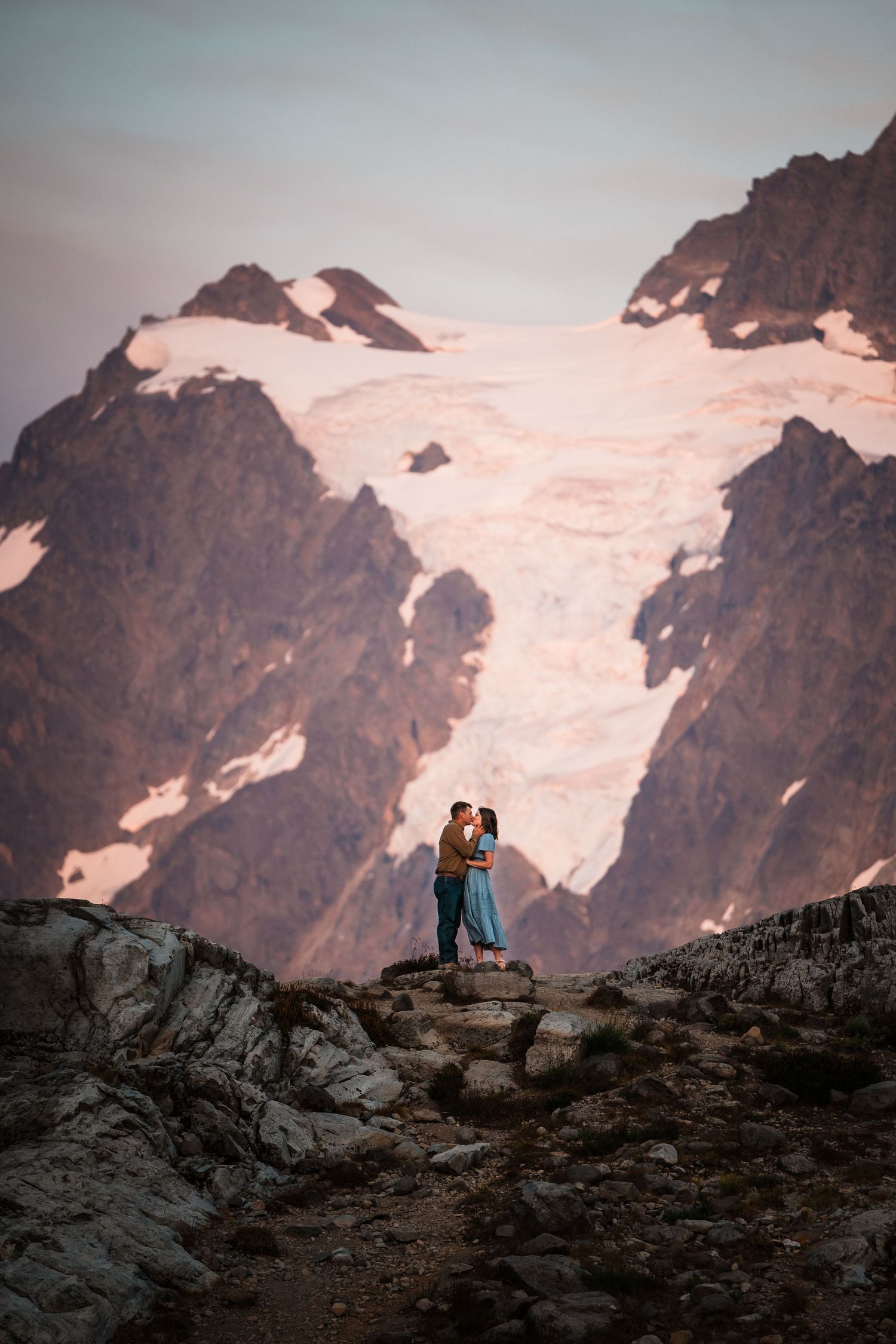 Two silhouettes sit on rocky mountain ridge against snowy peaks and pink sunset sky in dramatic alpine landscape.