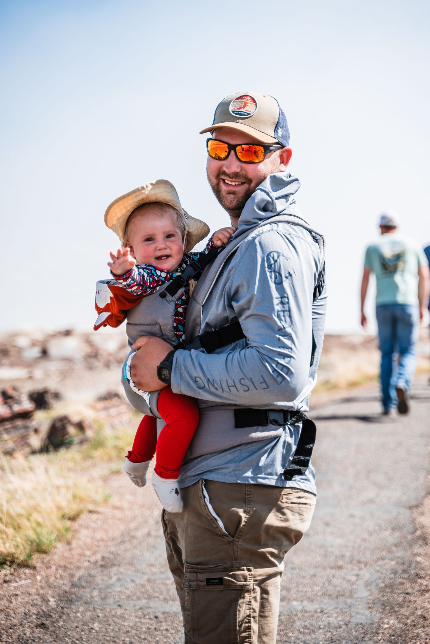 Two hikers on an outdoor trail enjoying a sunny day, one carrying a baby in a carrier against a blue sky background.