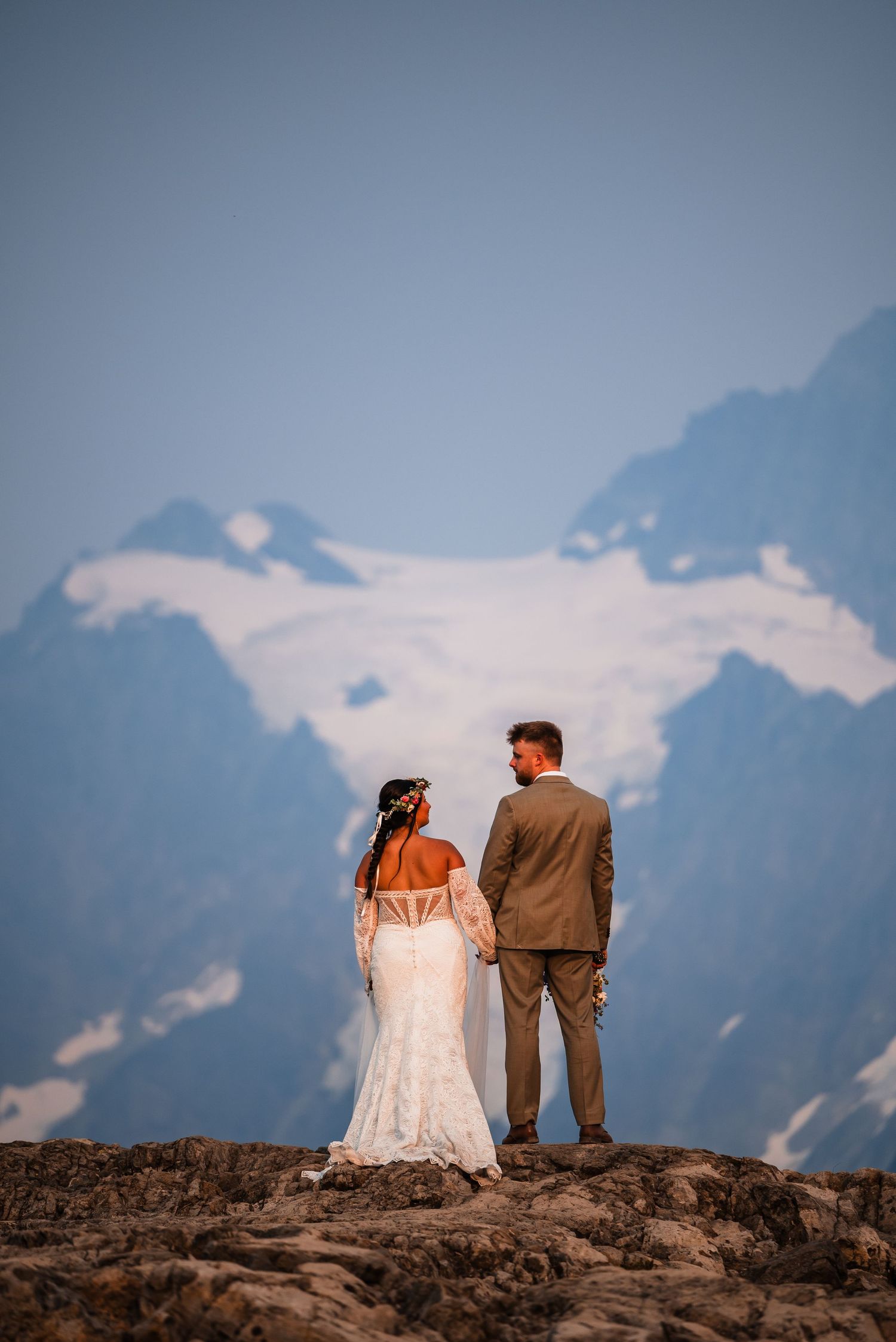 Silhouettes of a couple stand on rocky terrain against a backdrop of majestic snow-covered mountain peaks.
