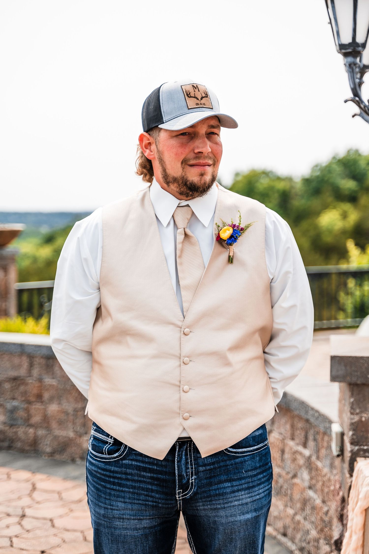 Groom wearing a beige vest and baseball cap standing against stone wall outdoors.
