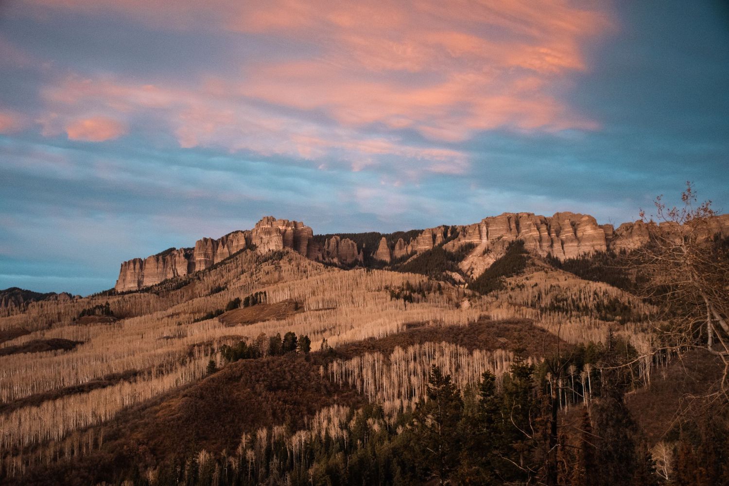 Rugged cliff face of mountain range captured at sunset with pink clouds and golden light illuminating the rocky terrain.