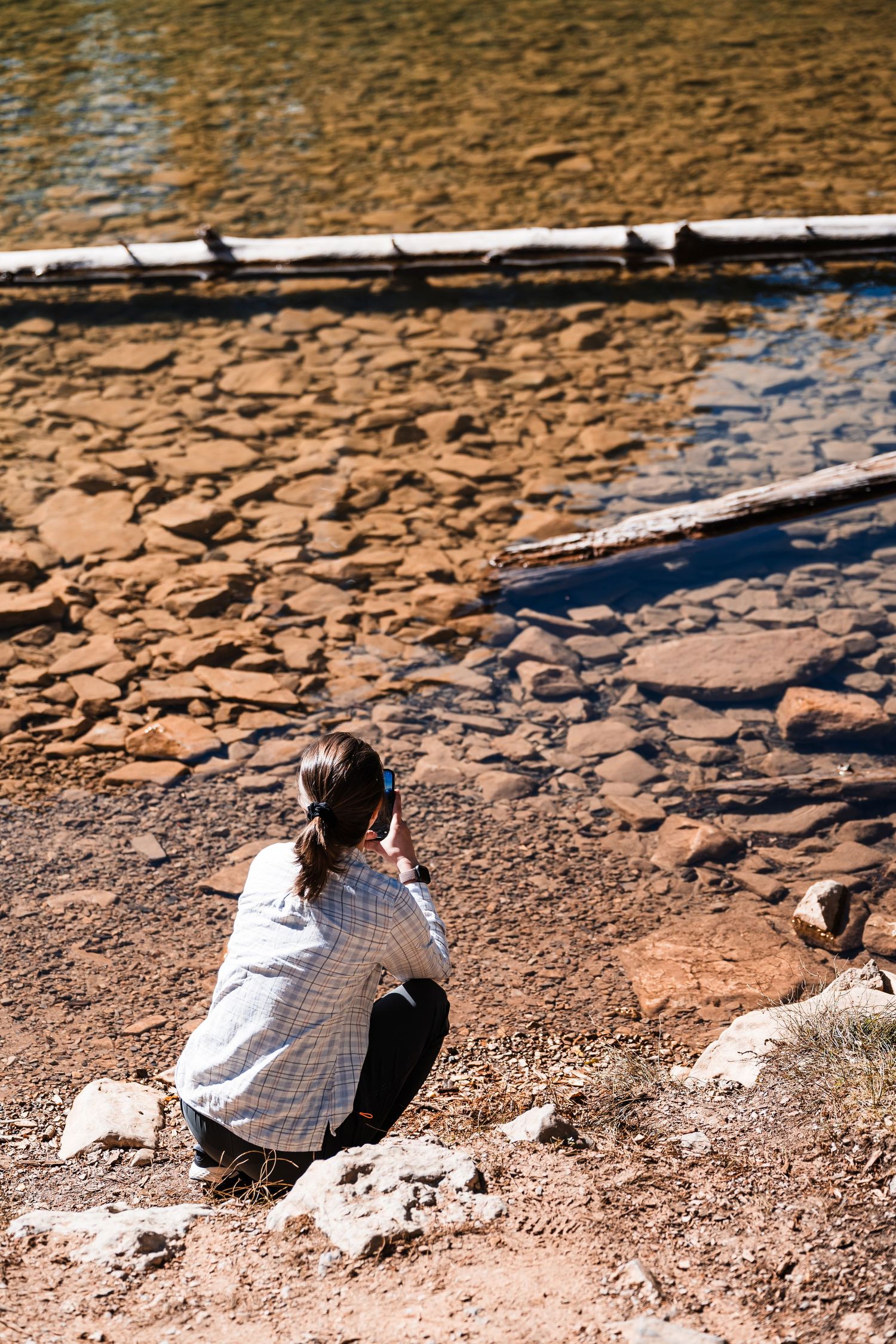 Person in white shirt sits on rocky riverbank photographing scenic water and stone landscape at sunset.