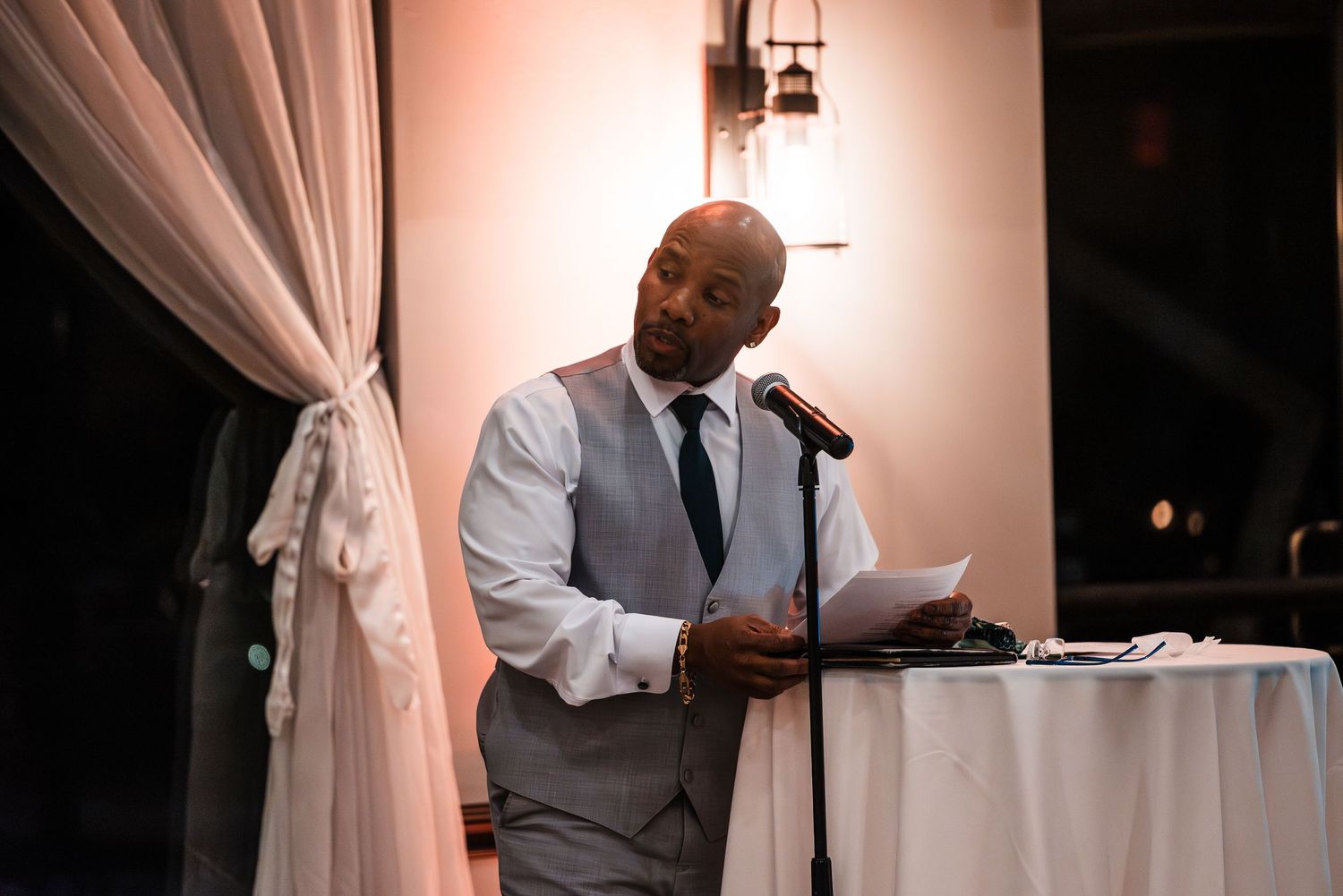 Man in grey vest delivers a heartfelt speech at a wedding reception podium with microphone.