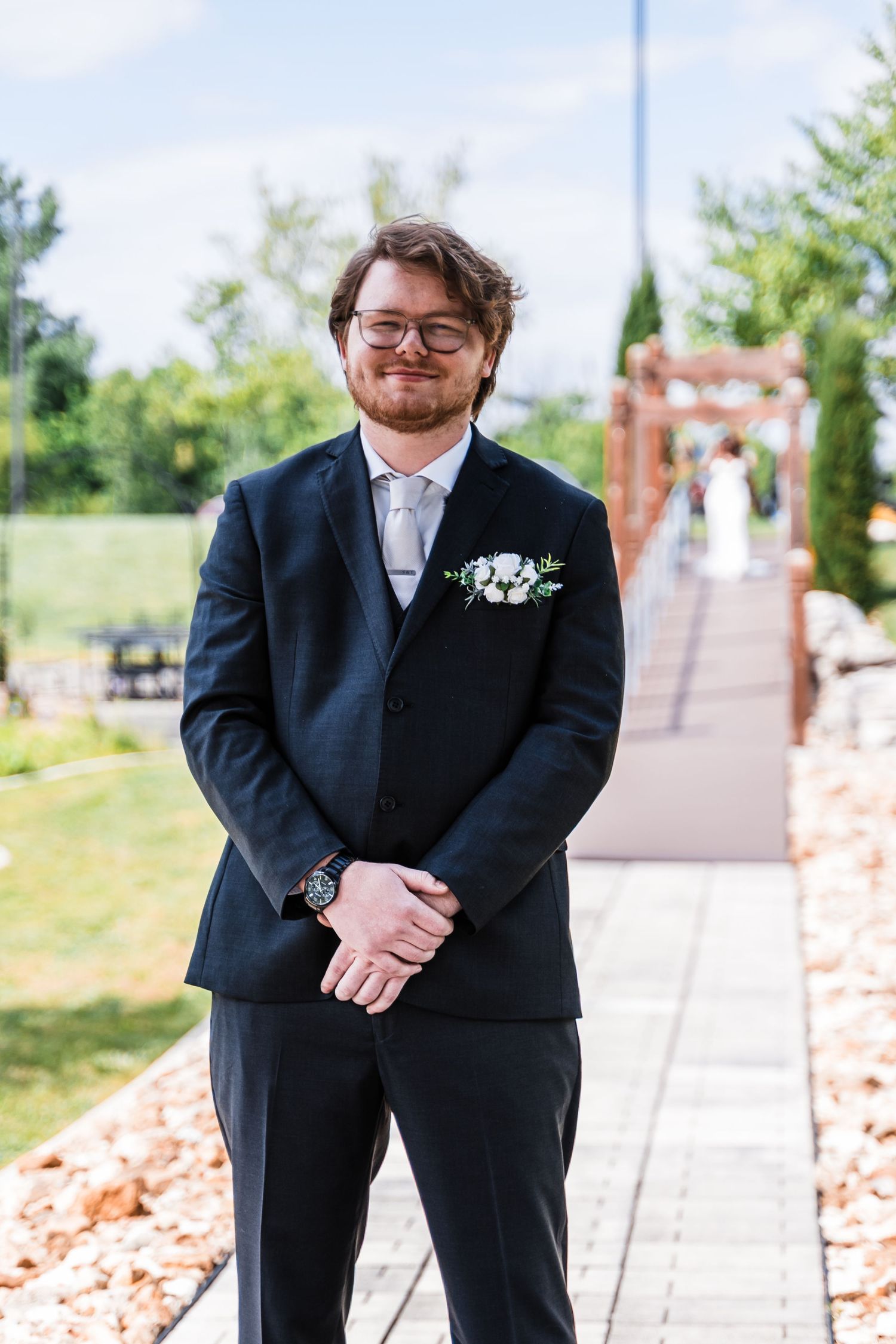 Smiling groom in dark suit with floral boutonniere standing outdoors on sunny wedding day.