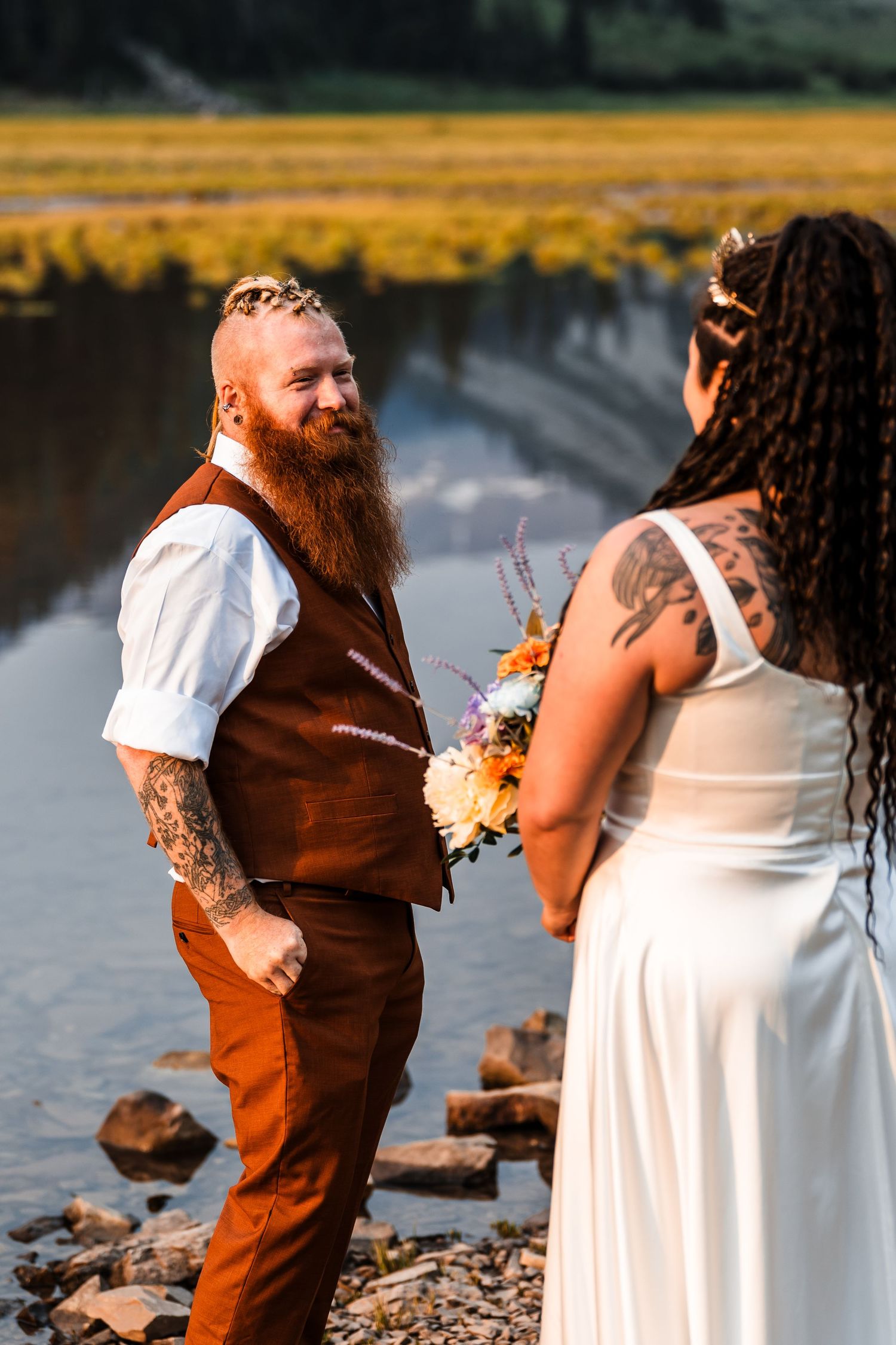 Bearded tattooed groom in rust suit smiling at bride holding colorful bouquet by mountain lake.