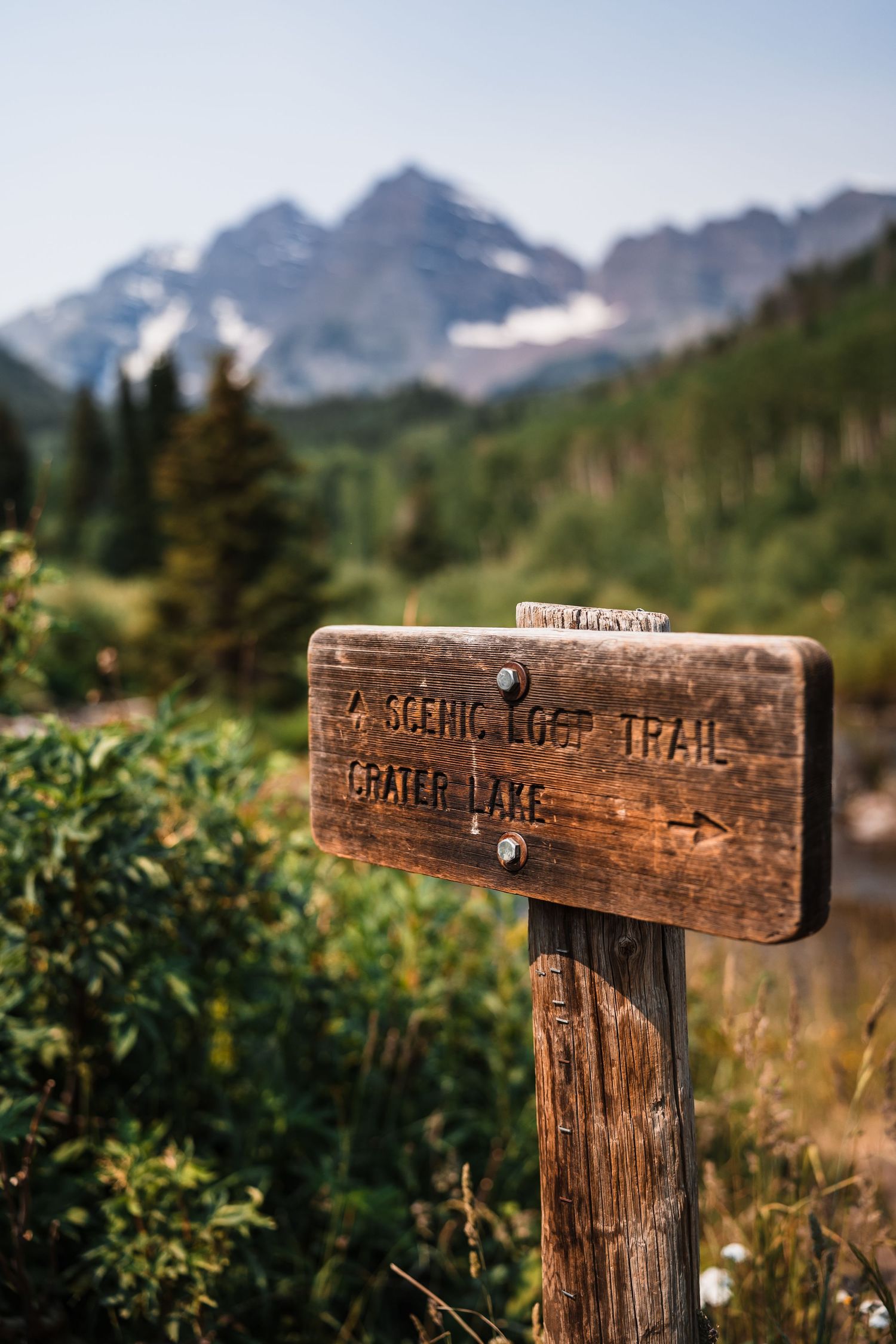 Wooden trail sign for Scenic Loop Trail and Crater Lake with snow-capped Maroon Bells mountains in background.