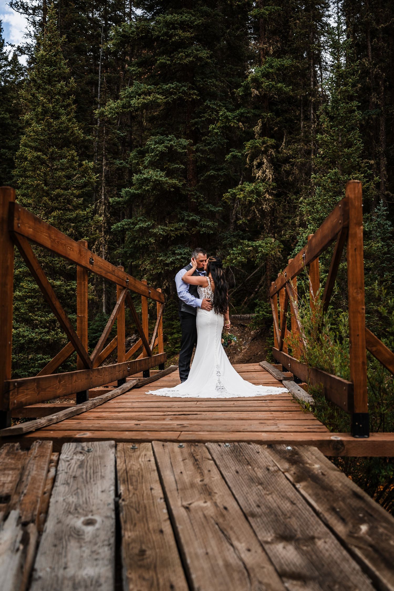 Bride and groom embrace romantically on a wooden bridge in a lush forest setting.