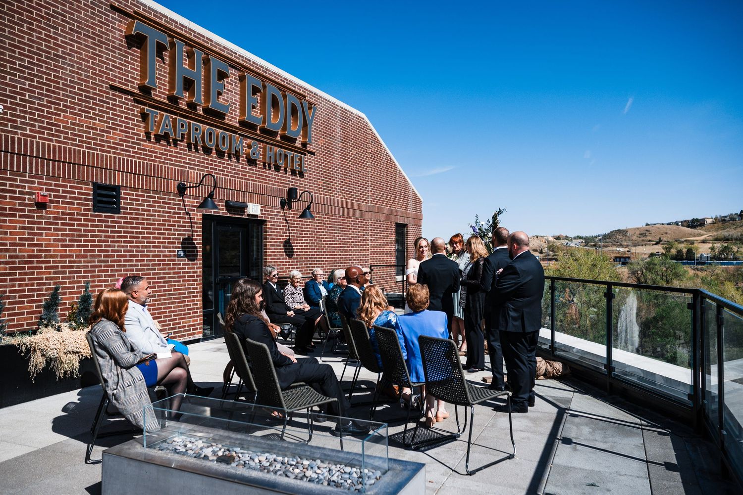 Wedding guests gathered on rooftop patio of The Eddy Taproom and Hotel under clear blue sky.