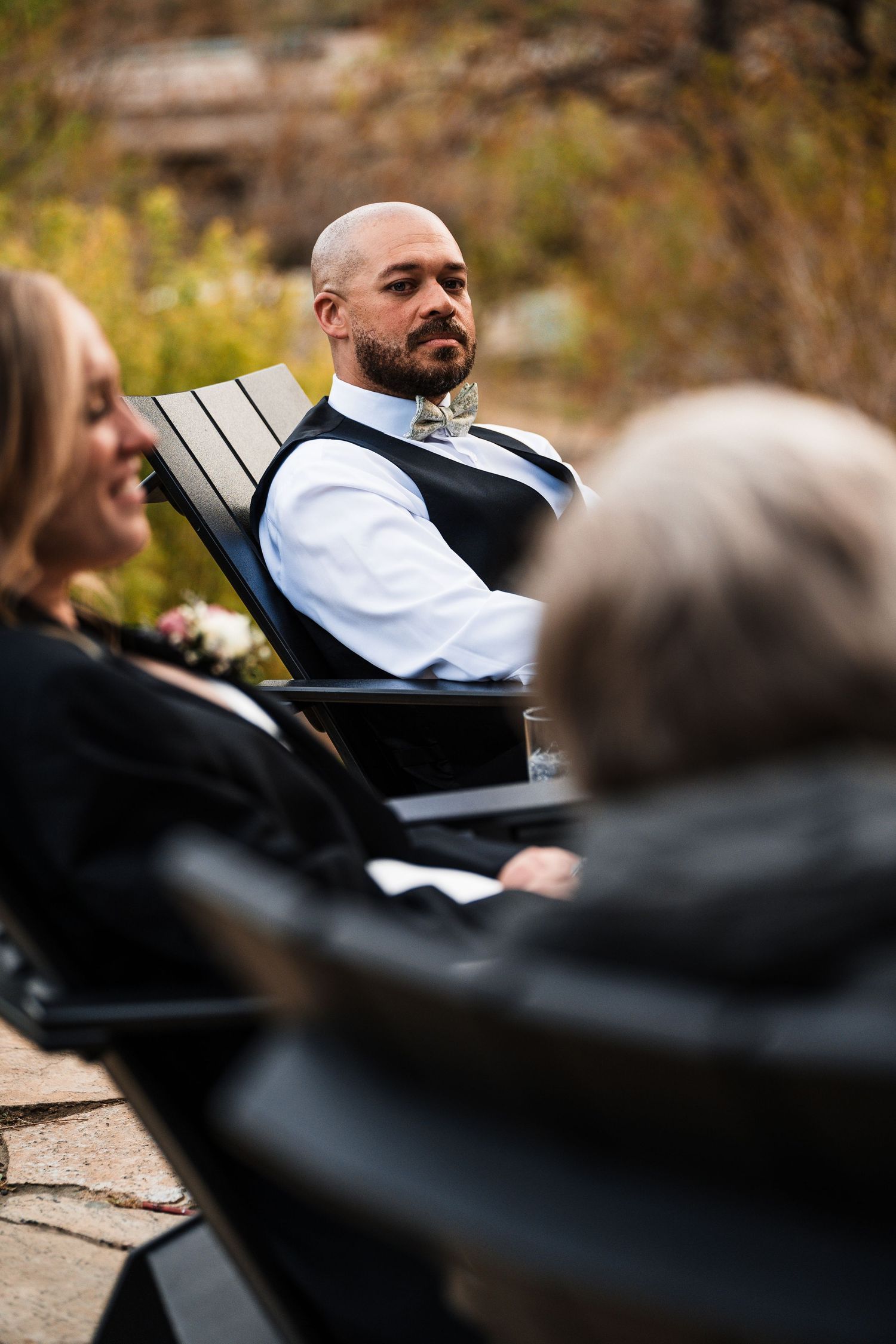 Bald groom with beard sits attentively at an outdoor fall wedding ceremony wearing a bow tie.